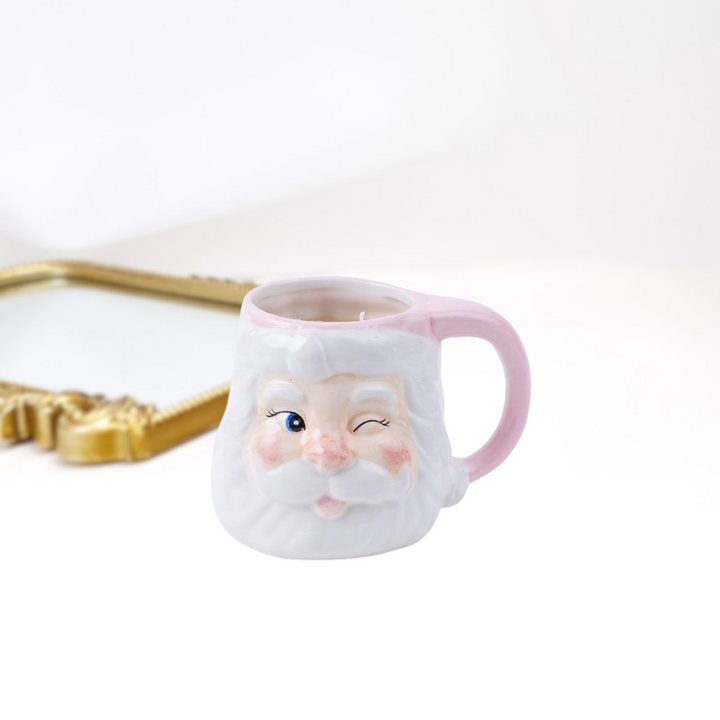 A gold mirror on a white background and a holiday santa mug candle in light pink.