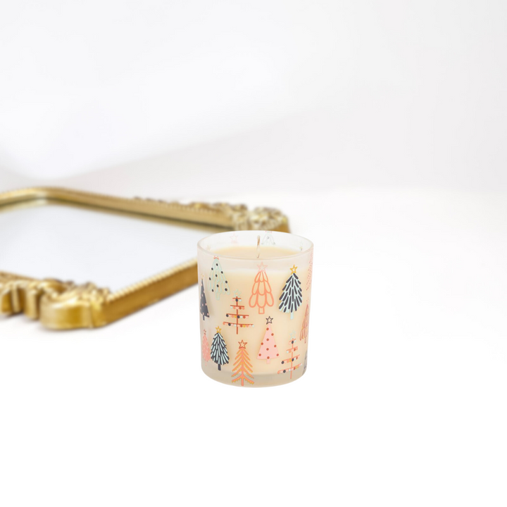 A gold mirror on a solid white background. A short clear candle with different colorful christmas trees on it.