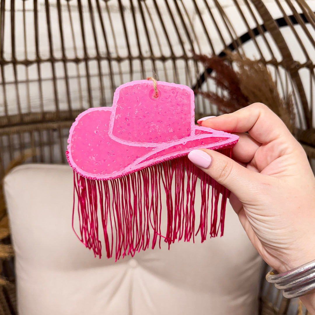 Hot pink hat car freshie with hot pink fringe hanging, hand holding freshie in front of wooden rustic background