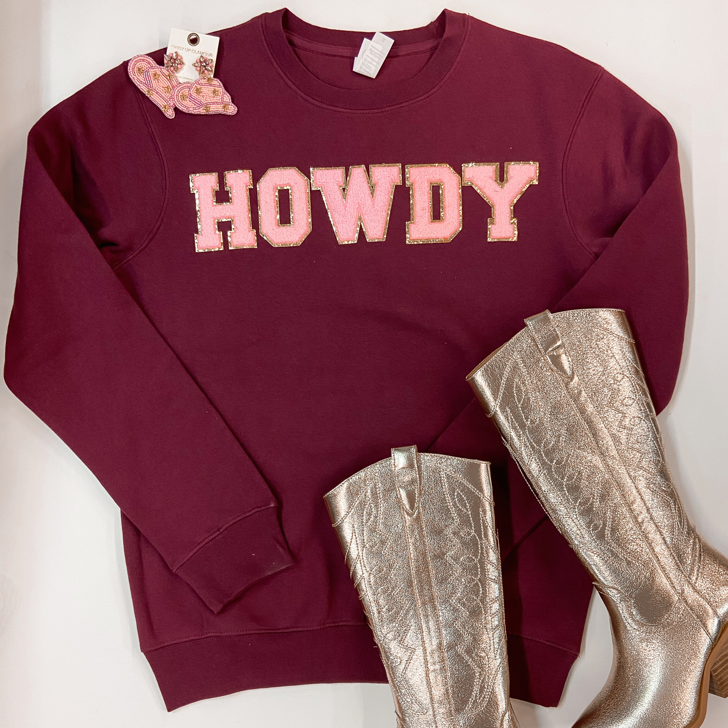 Picture features a maroon, pink, and gold Howdy Chenille sweatshirt paired with gold boots and some pink cowboy hat earrings.