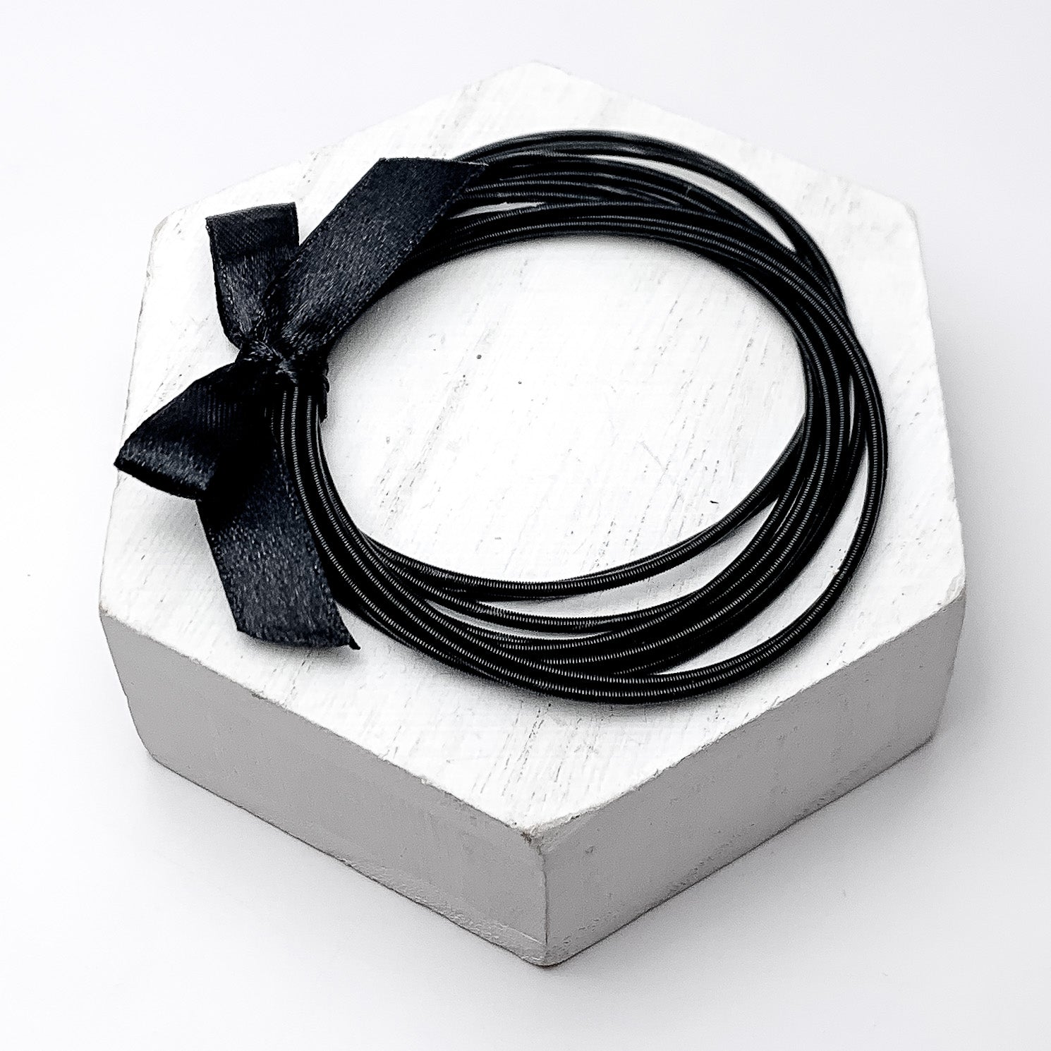 Guitar String Bracelets with Bow in Jet Black. Pictured on a white background sitting on top of a white block.