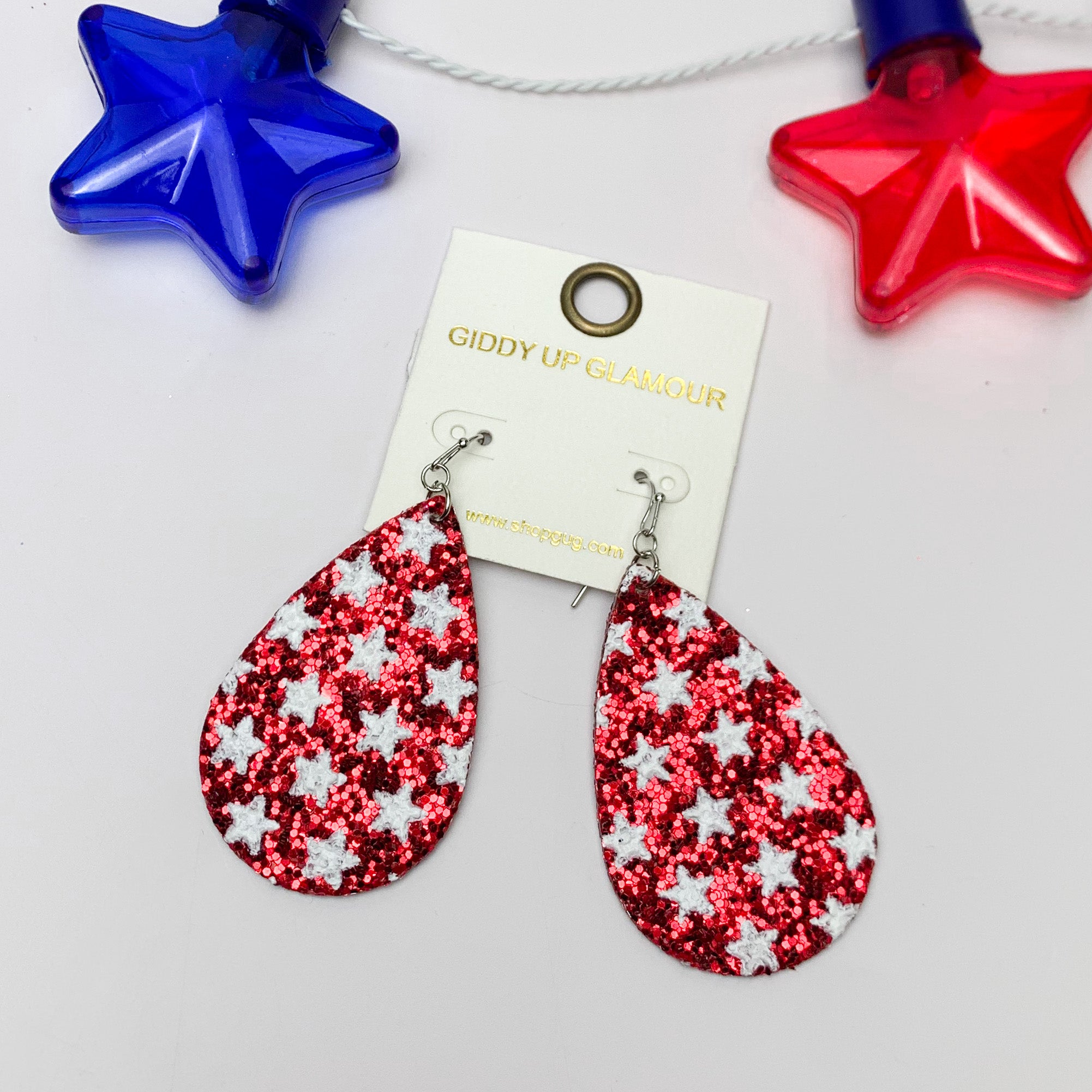 Festive Sparkly Red Drop Earrings Filled With White Stars. Pictured on a white background with red and blue stars at the top as an accessory. 
