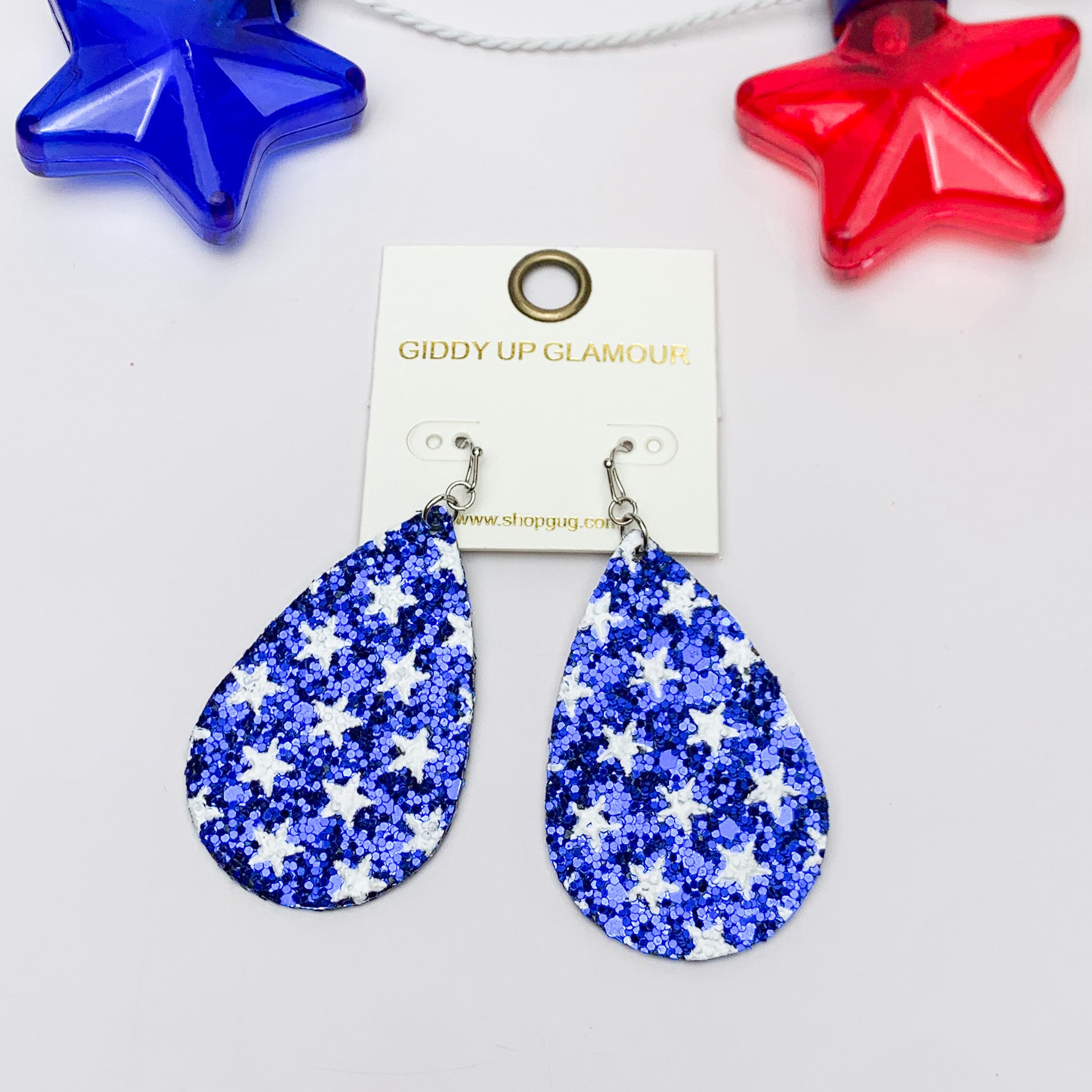Festive Sparkly Blue Drop Earrings Filled With White Stars. Pictured on a white background with a red and blue star behind the earrings.