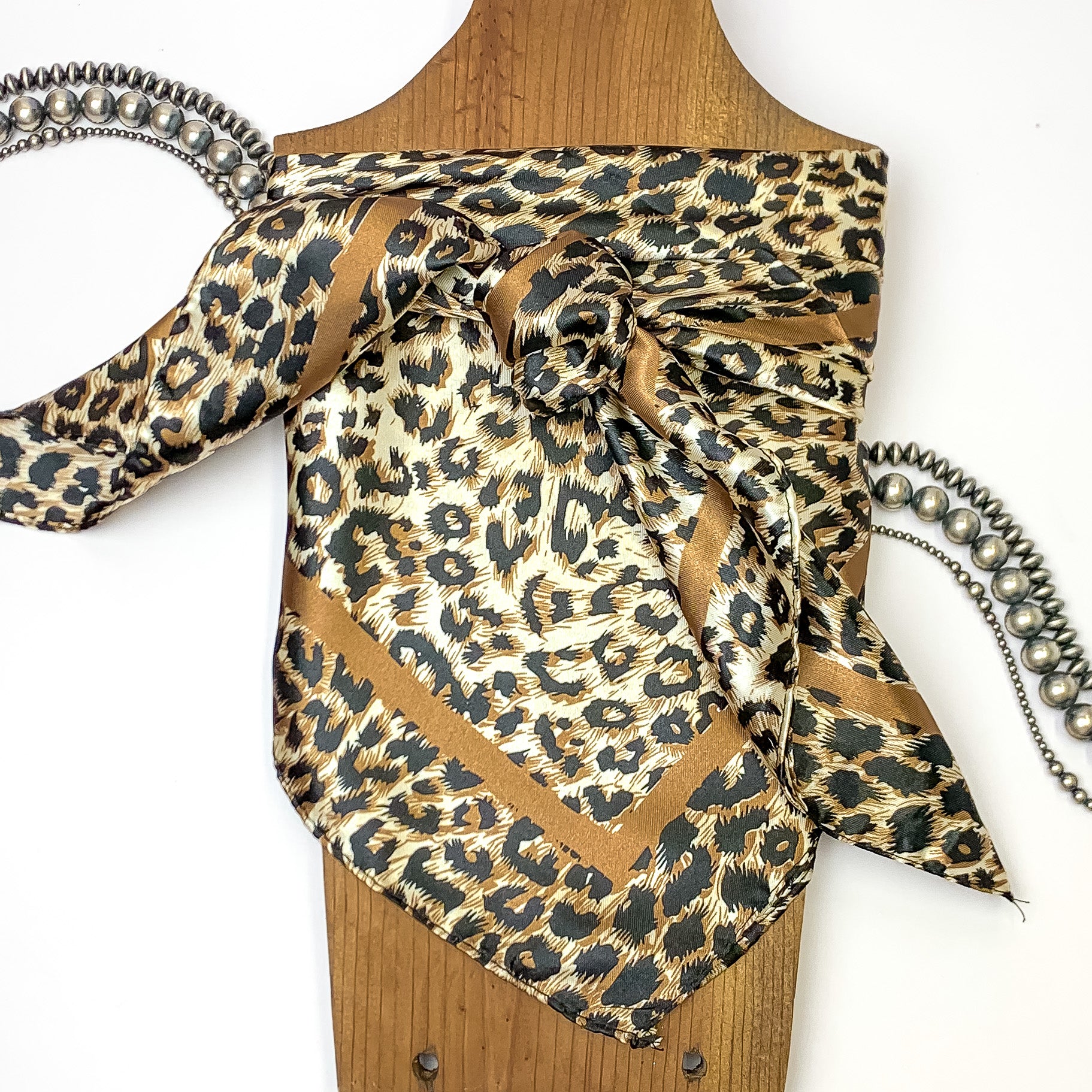 Leopard print scarf tied around a wooden necklace holder, pictured on a white background that has silver beads across. 