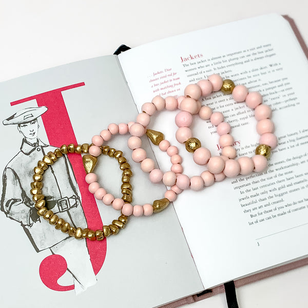 Set of Four | Stretchy Light Pink Beaded Bracelets featuring a Gold Tone Bracelet. Pictured on a white background. Bracelets are laying on top of an open book.