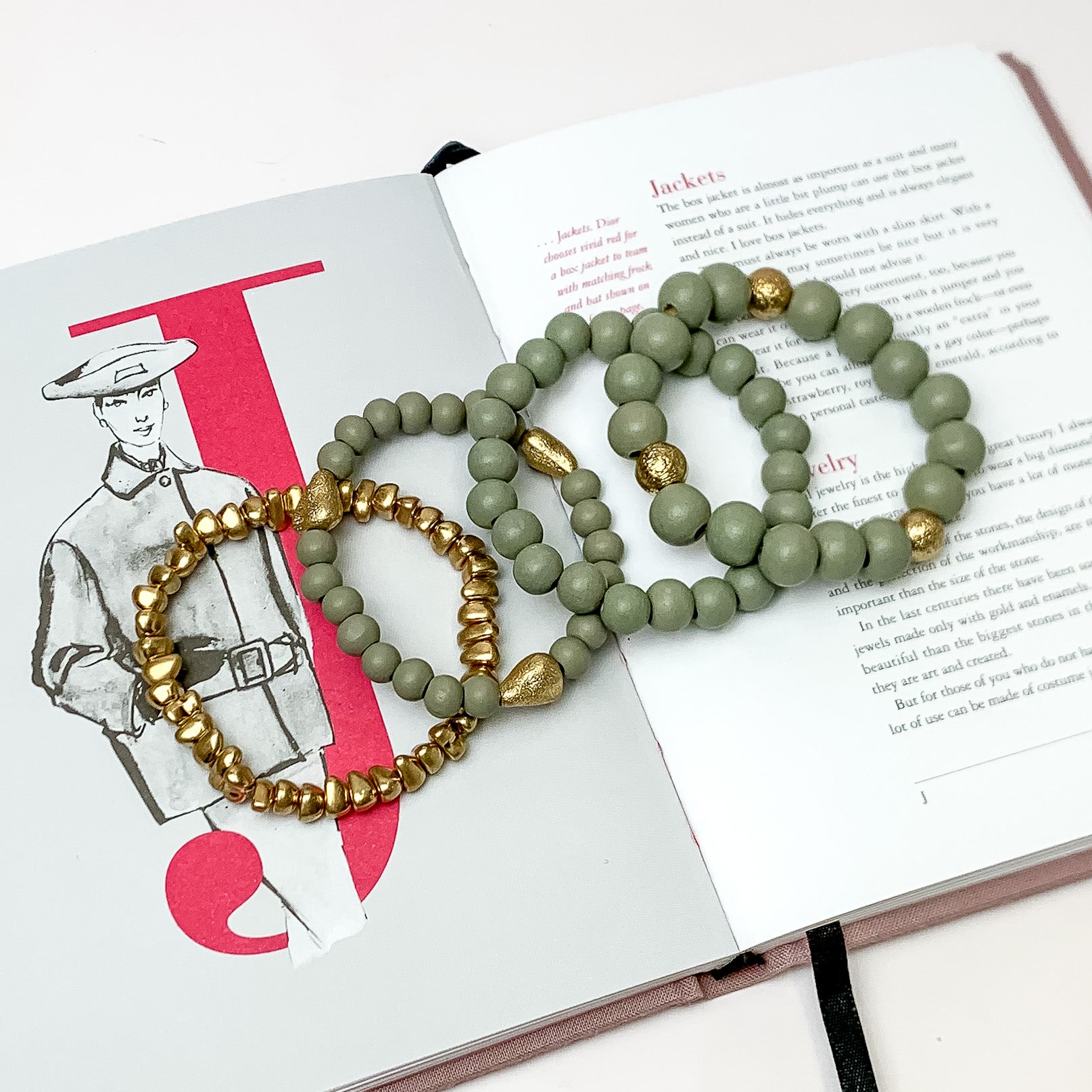 Set of Four | Stretchy Grey Beaded Bracelets featuring a Gold Tone Bracelet. Pictured on a white background. Bracelets are laying on top of an open book.