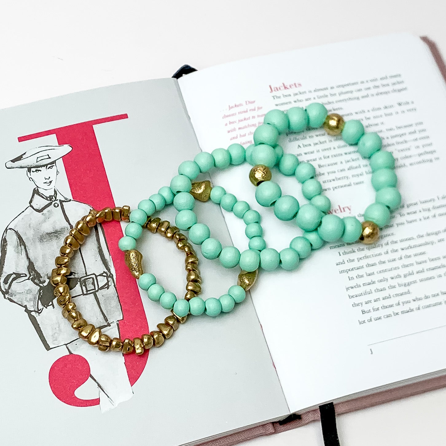 Set of Four | Stretchy Turquoise Blue Beaded Bracelets featuring a Gold Tone Bracelet. Pictured on a white background. Bracelets are laying on top of an open book.