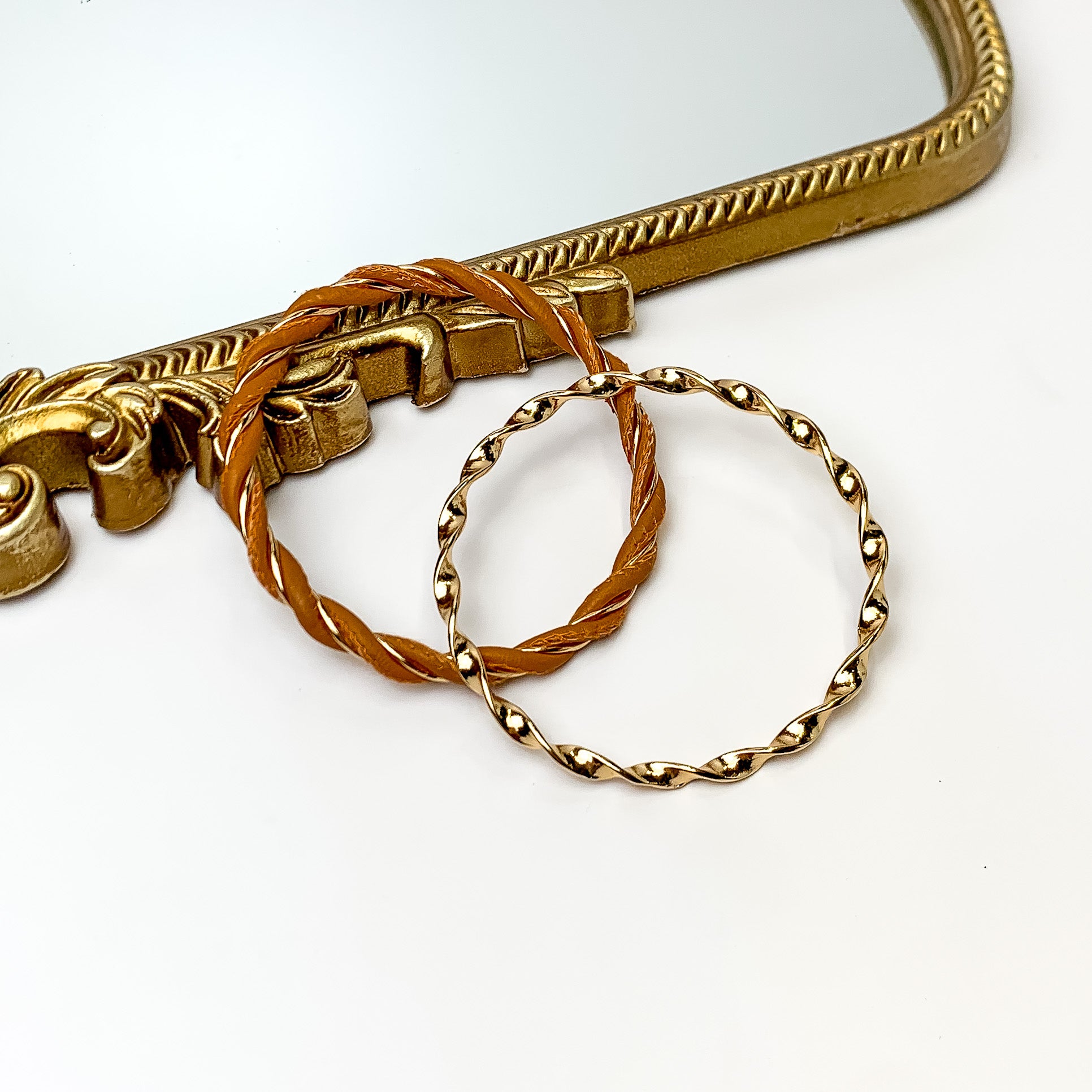 Pictured is a gold twist bangle and gold and tan twist bangle. These bangles are pictured partially leaning on a gold mirror on a white background. 