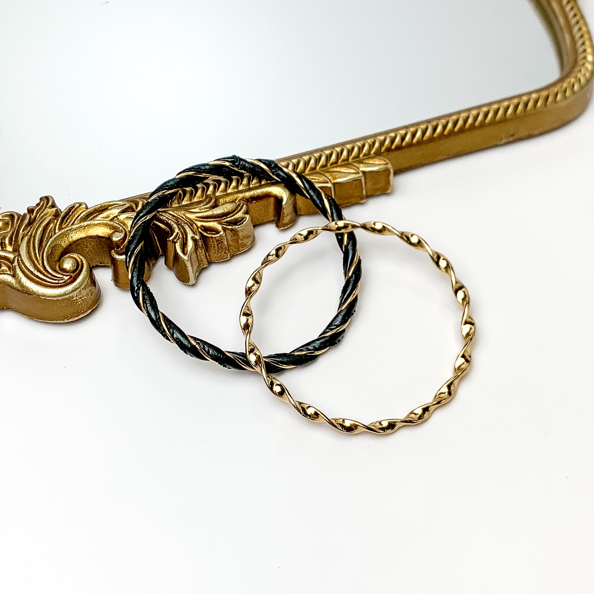 Pictured is a gold twist bangle and gold and black twist bangle. These bangles are pictured partially leaning on a gold mirror on a white background. 