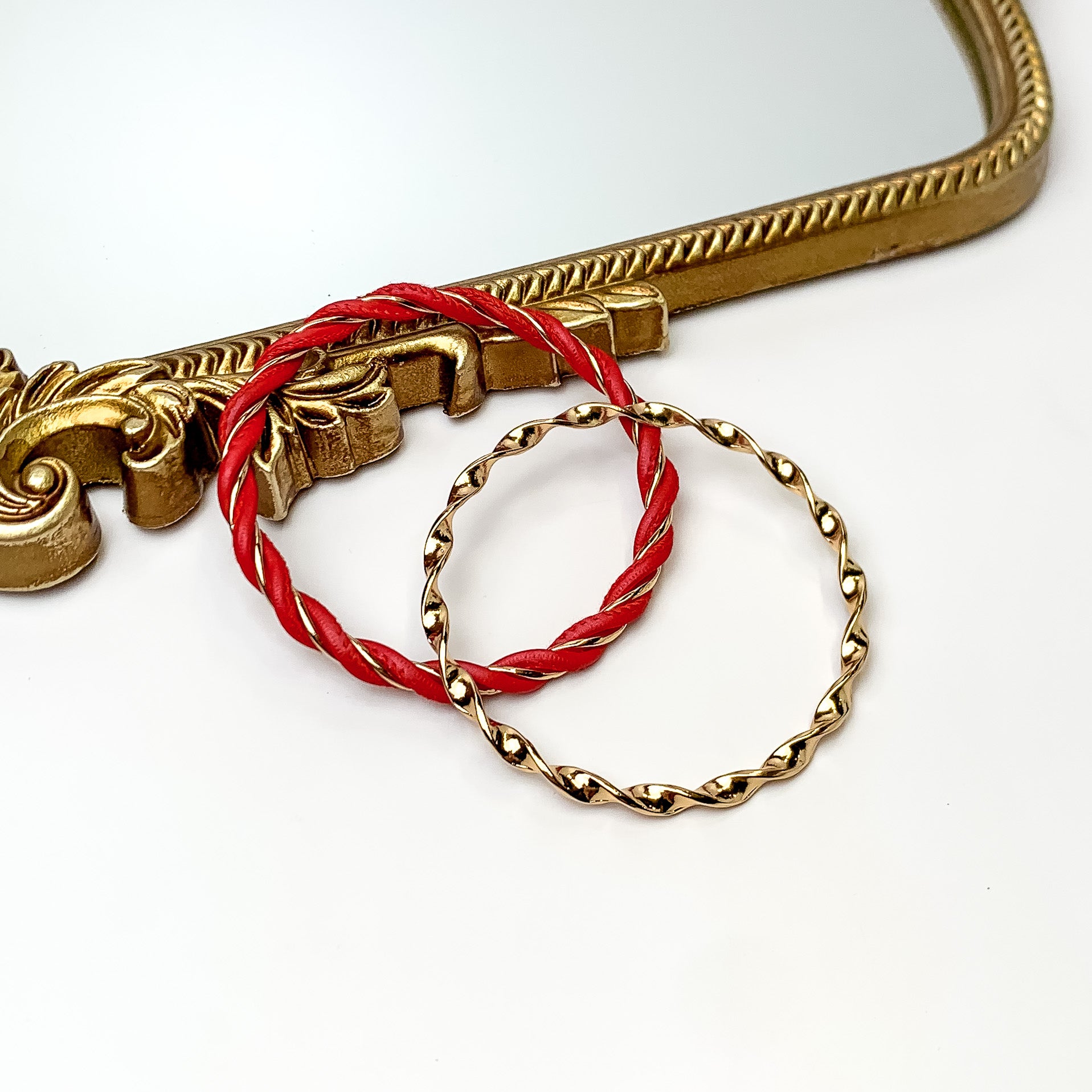 Pictured is a gold twist bangle and gold and red twist bangle. These bangles are pictured partially leaning on a gold mirror on a white background. 