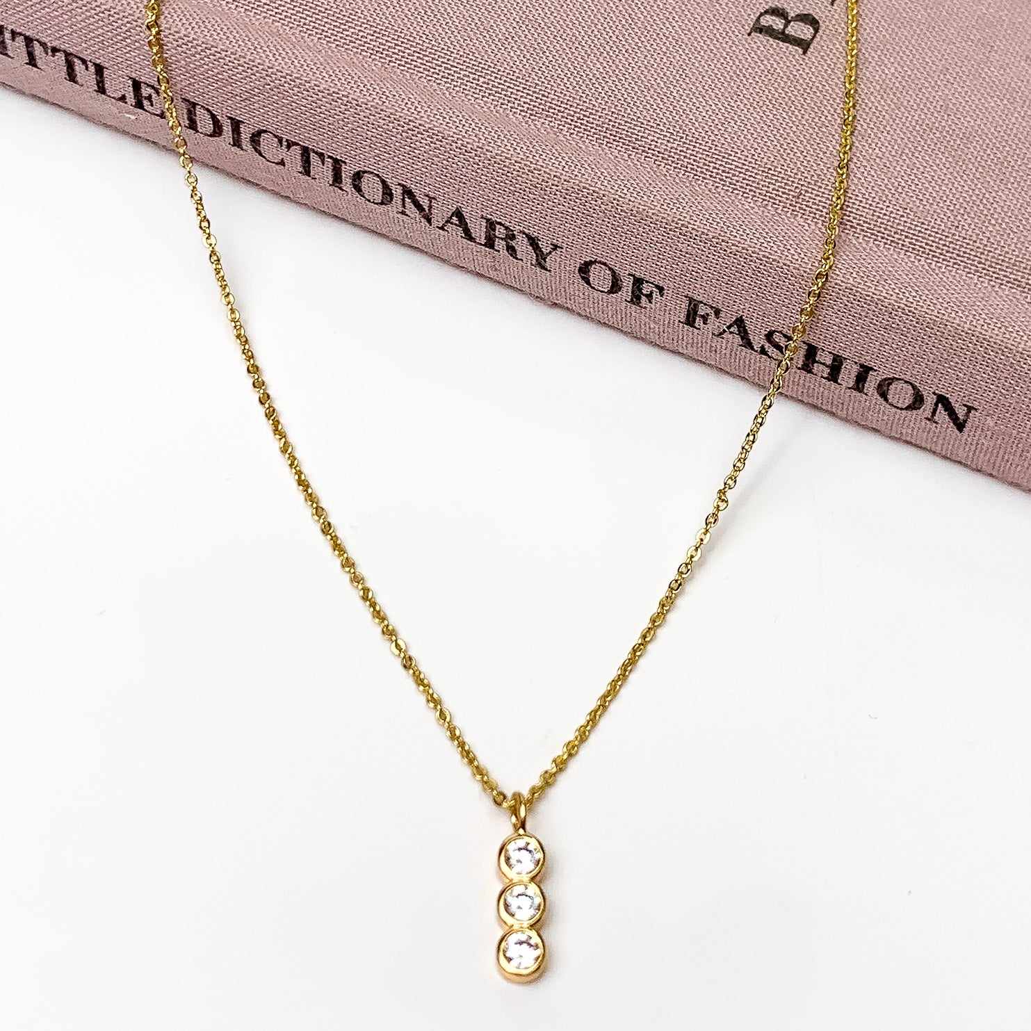 Gold Tone Necklace with Triple Clear Crystal Pendant. Pictured on a white background with the necklace laying on a book.