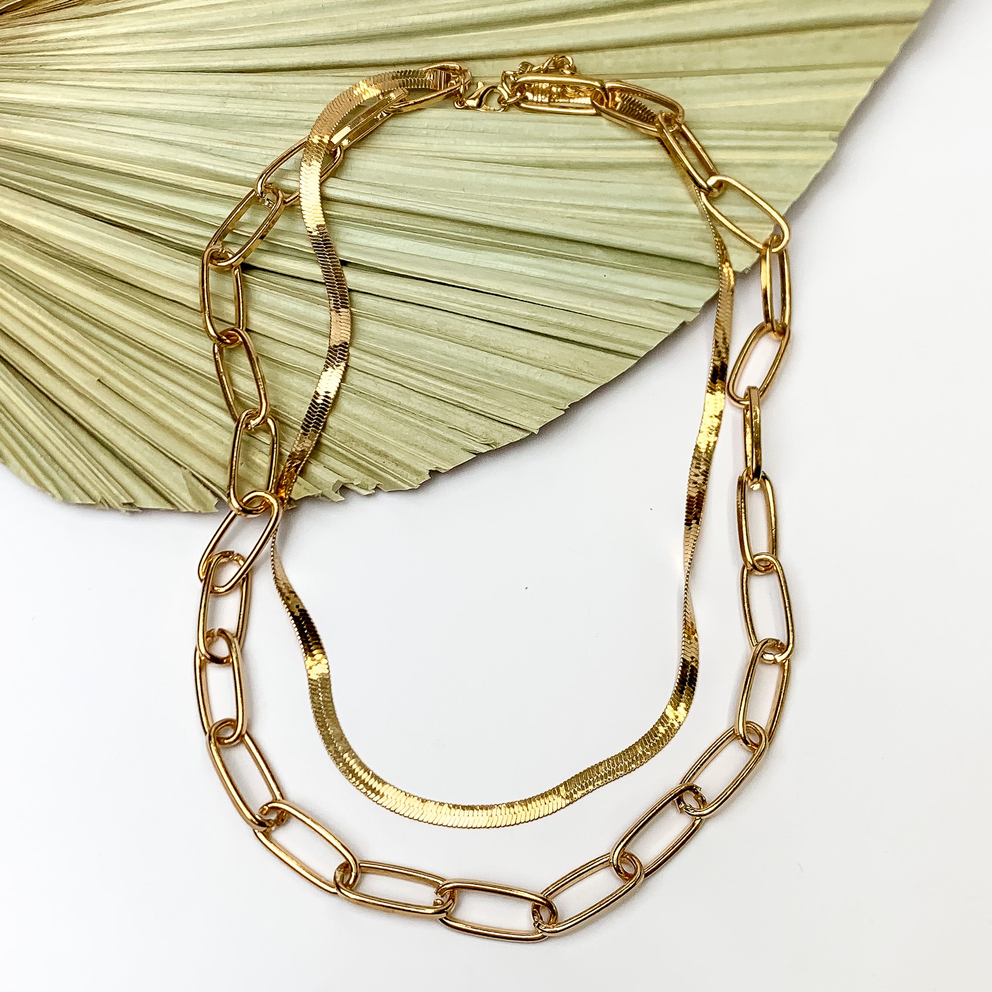 Gold Tone Double Layered Chain Necklace. Pictured on a white background with the necklace laying on a leaf fan.