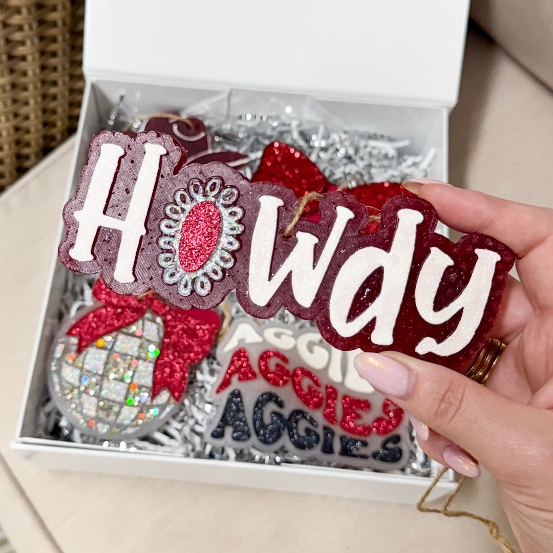 A hand holding a maroon car freshie with 'Howdy' written in glittery white lettering