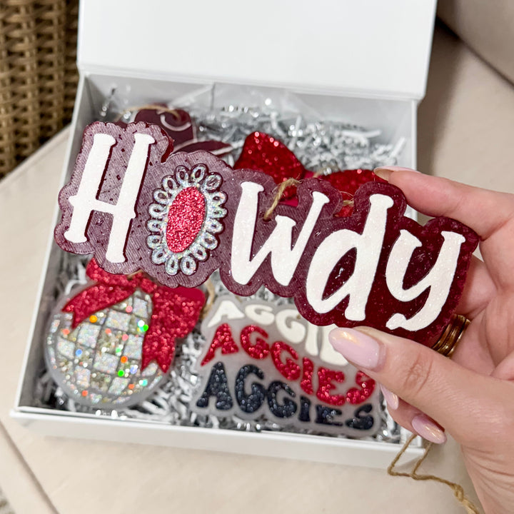 A hand holding a maroon car freshie with 'Howdy' written in glittery white lettering
