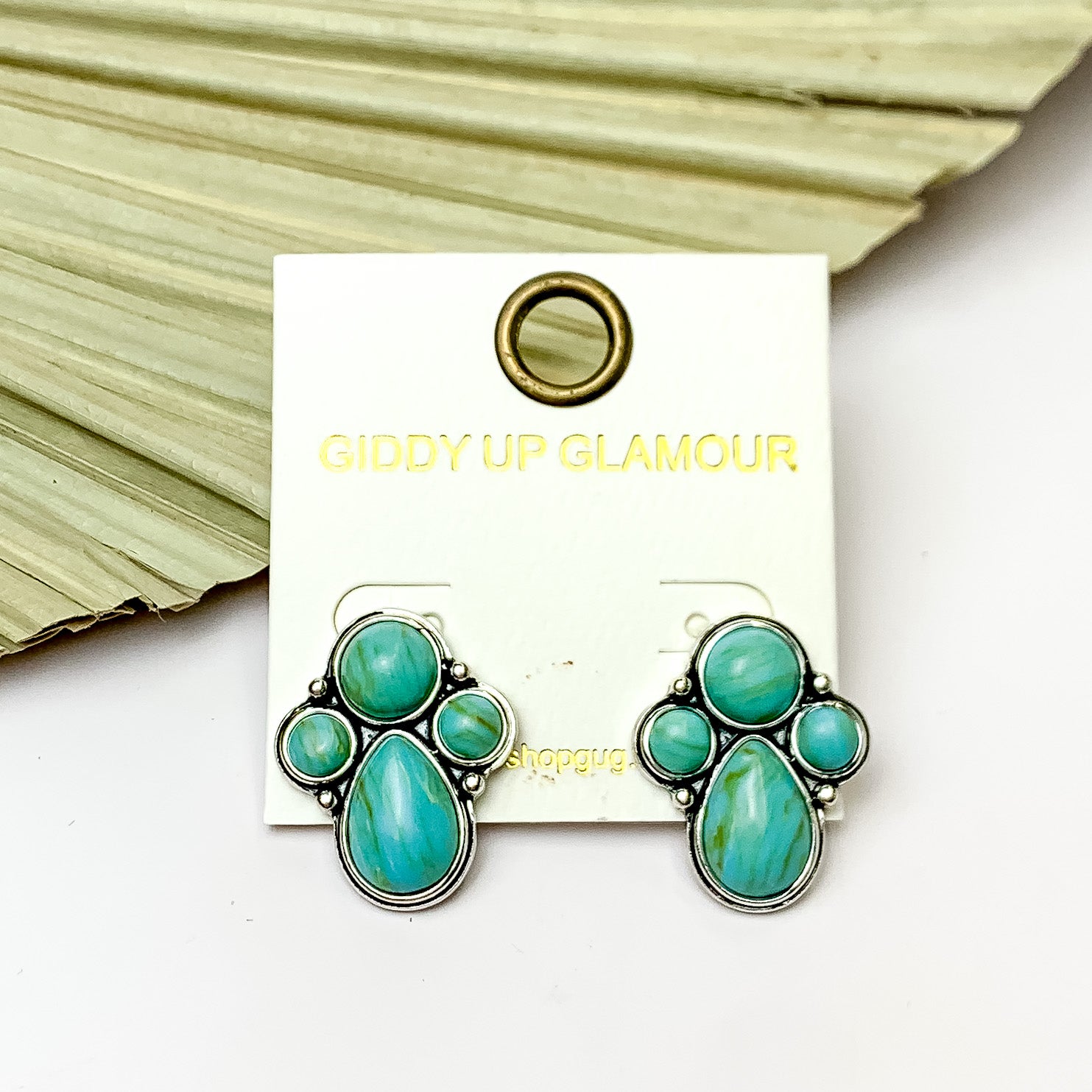 Silver Tone Cluster Stone Earrings in Turquoise Green. Pictured on a white background with a leaf fan behind the earrings. 