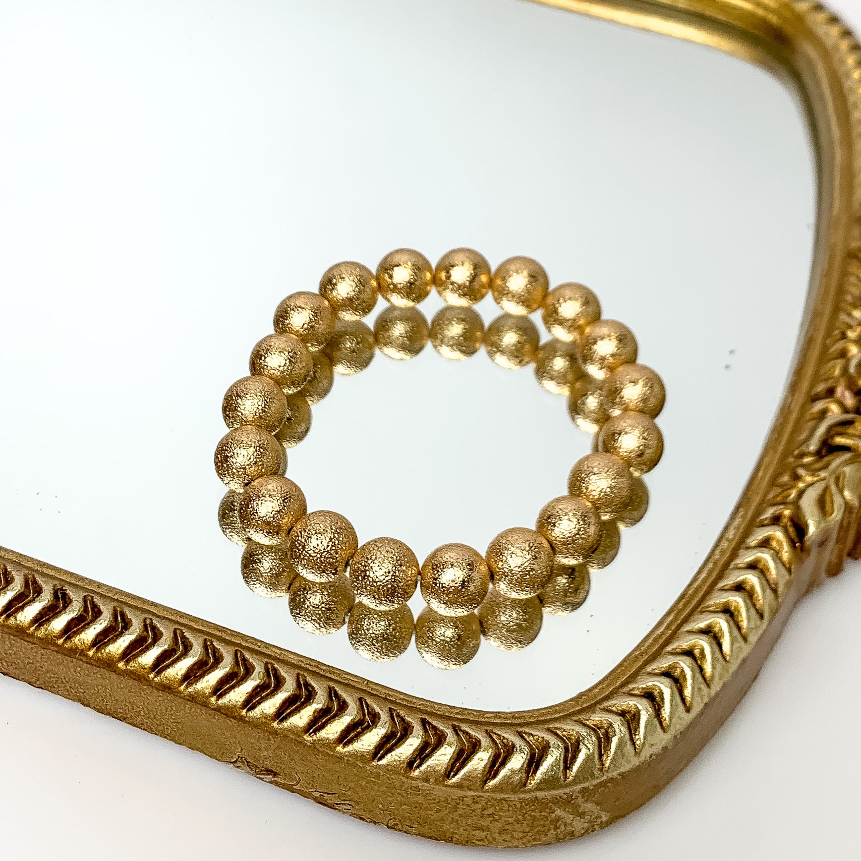 Gold beaded bracelet pictured on a gold mirror on a white background. 