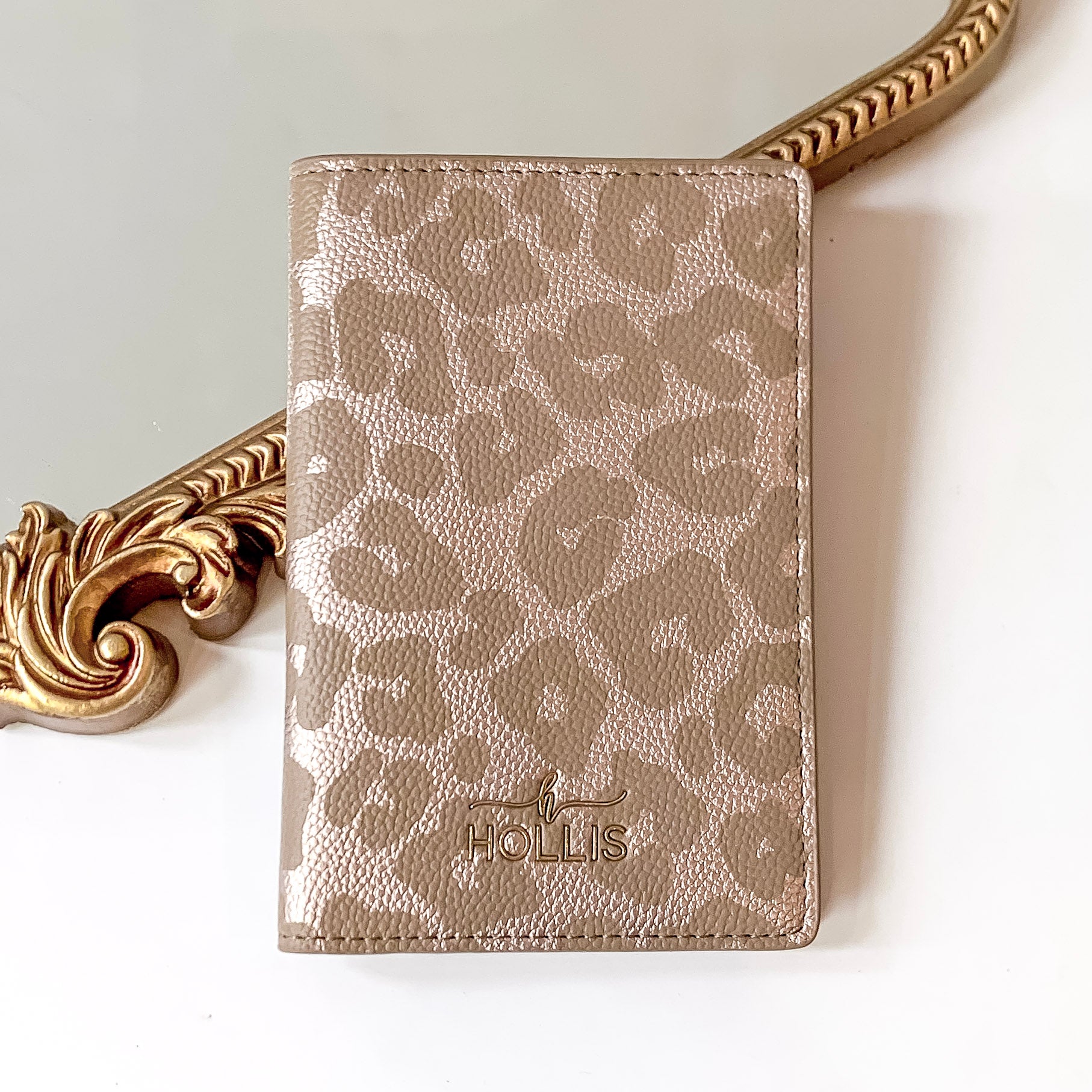 Leopard print passport holder with a gold HOLLIS emblem at the bottom. This is pictured laying partially on a gold mirror on a white background.