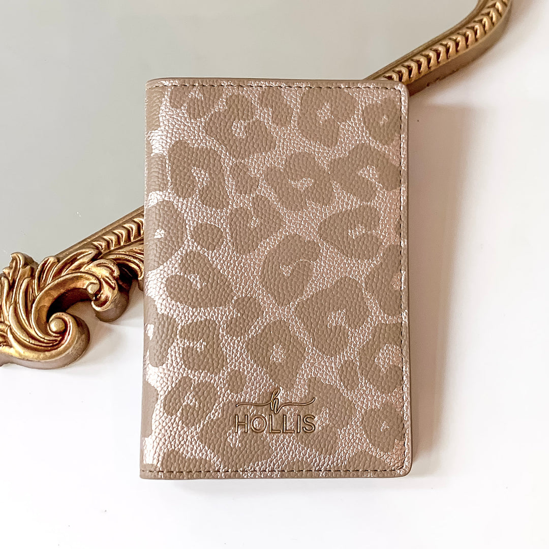 Leopard print passport holder with a gold HOLLIS emblem at the bottom. This is pictured laying partially on a gold mirror on a white background.