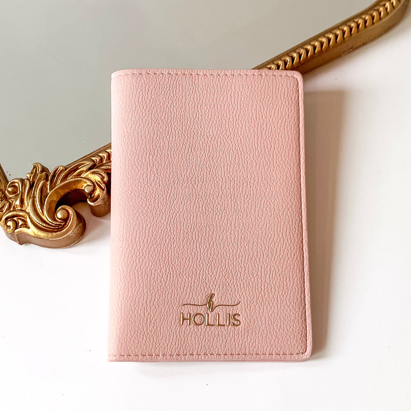 Blush pink passport holder with a gold HOLLIS emblem at the bottom. This is pictured laying partially on a gold mirror on a white background. 