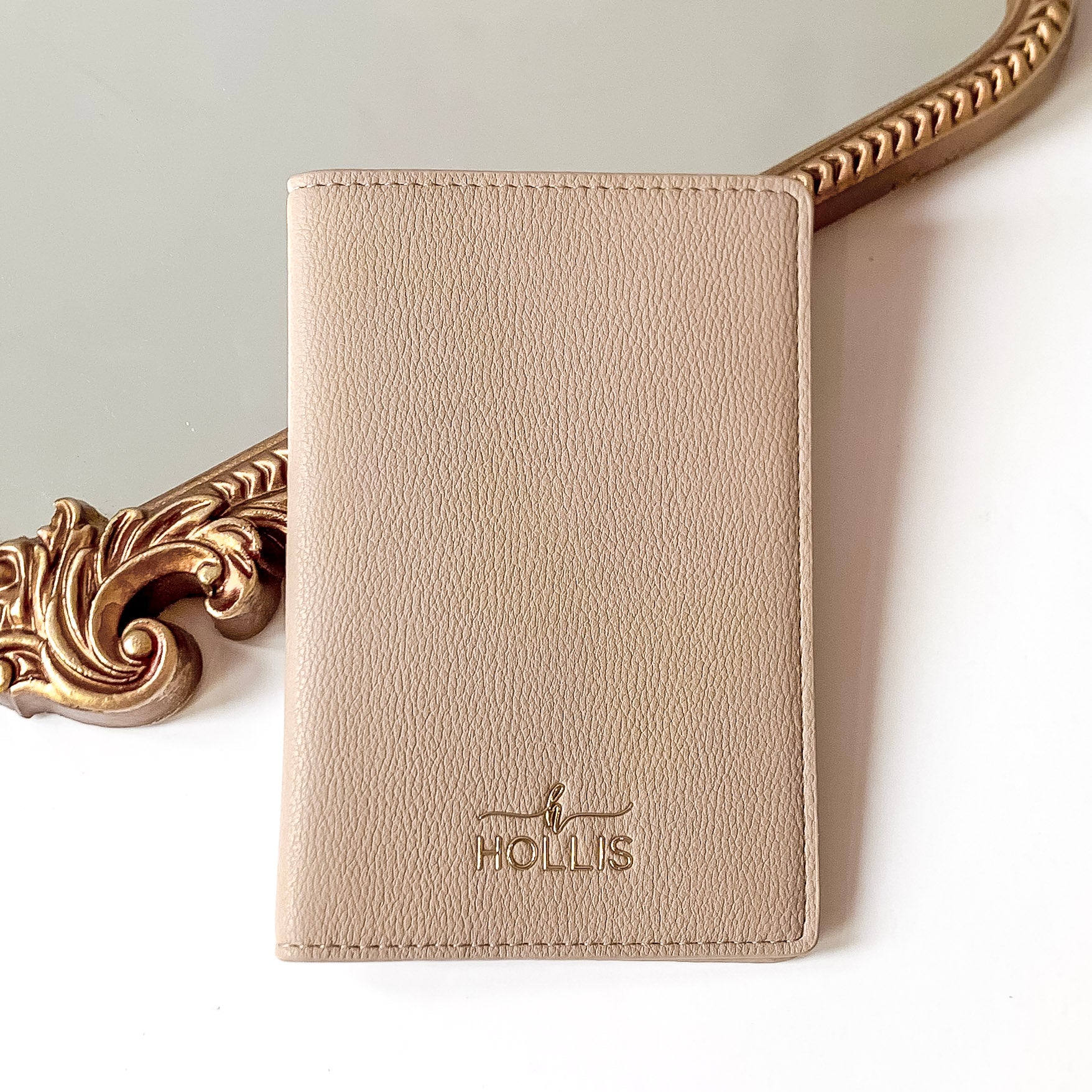 Nude passport holder with a gold HOLLIS emblem at the bottom. This is pictured laying partially on a gold mirror on a white background.