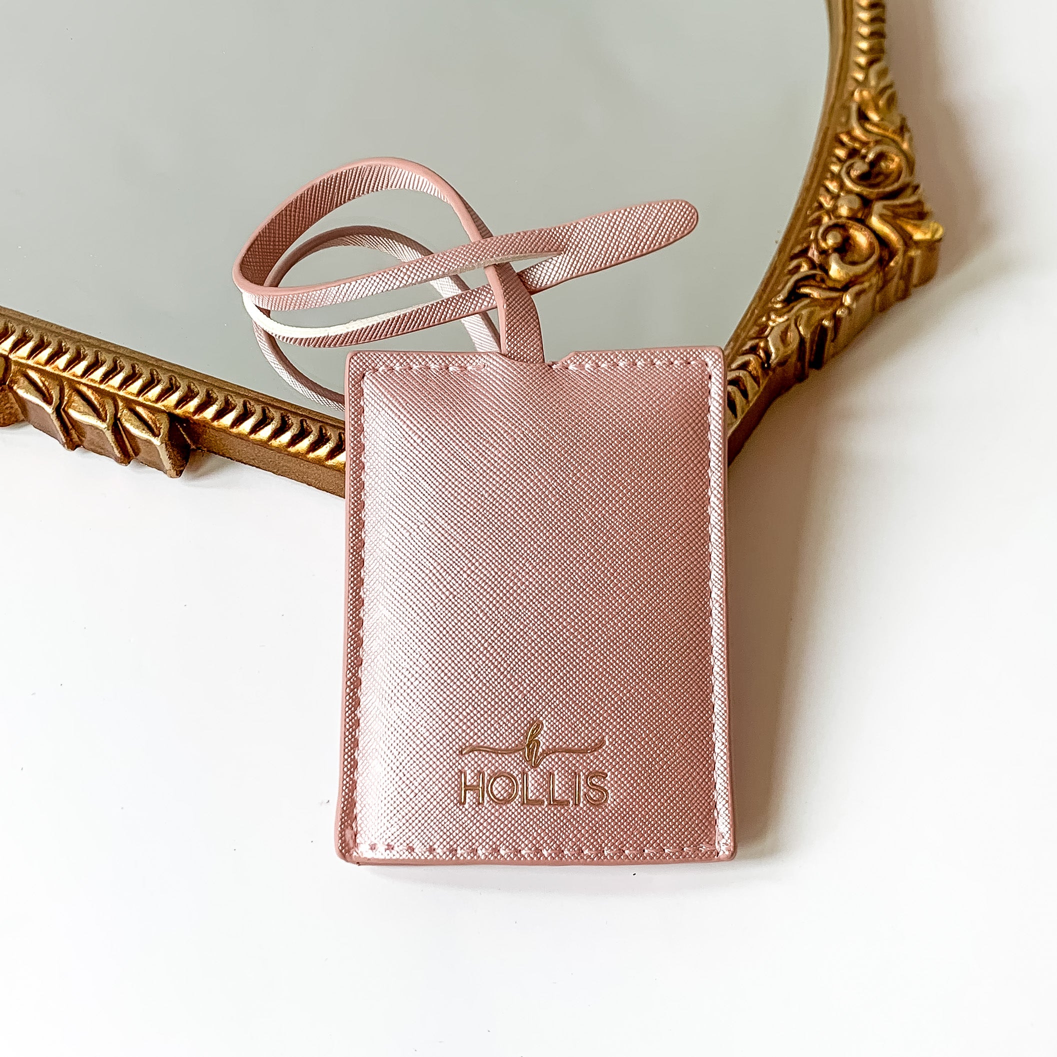 Metallic blush pink luggage tag with a gold HOLLIS emblem at the bottom. This luggage tag is pictured partially laying on a gold mirror on a white background. 