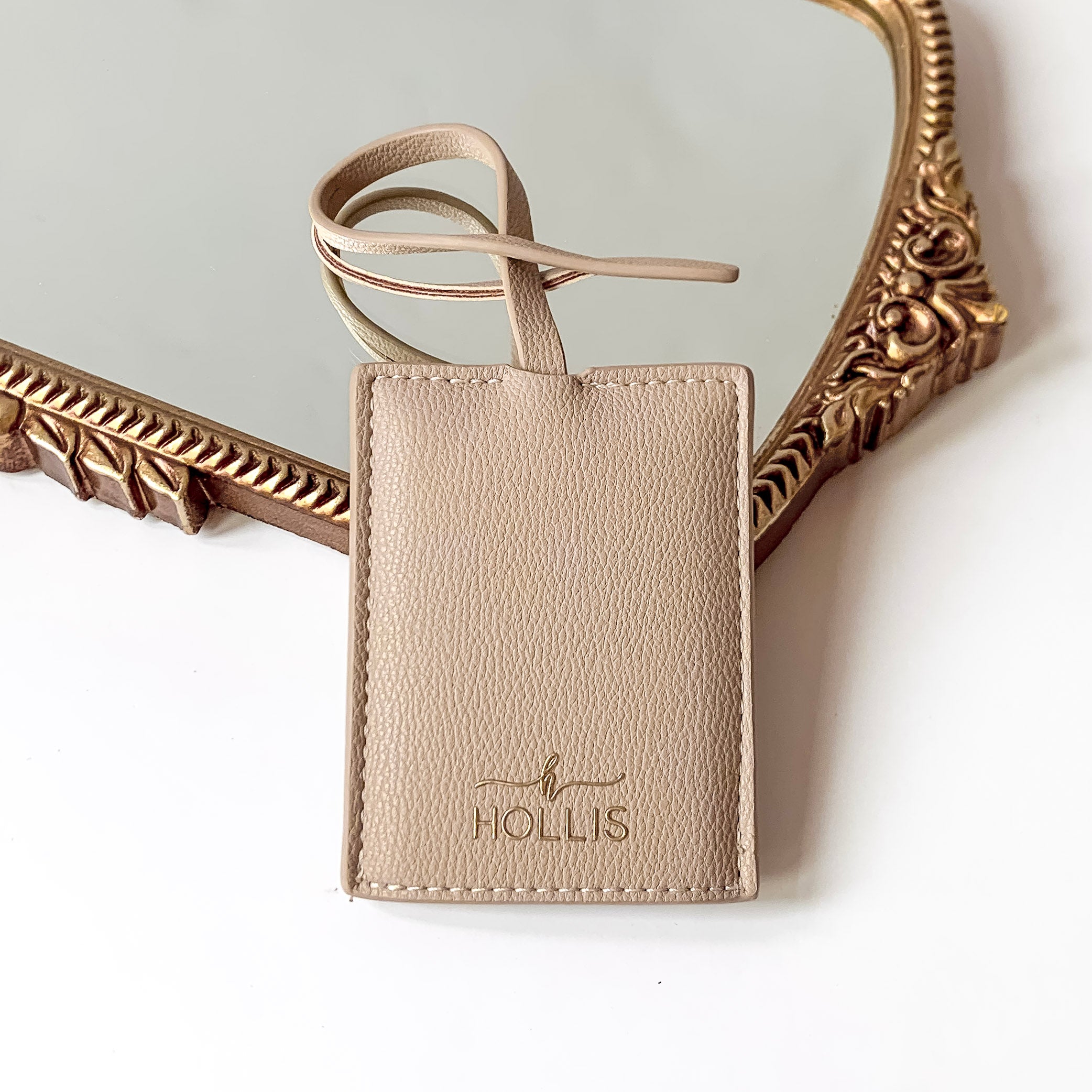 Nude luggage tag with a gold HOLLIS emblem at the bottom. This is pictured laying partially on a gold mirror on a white background.