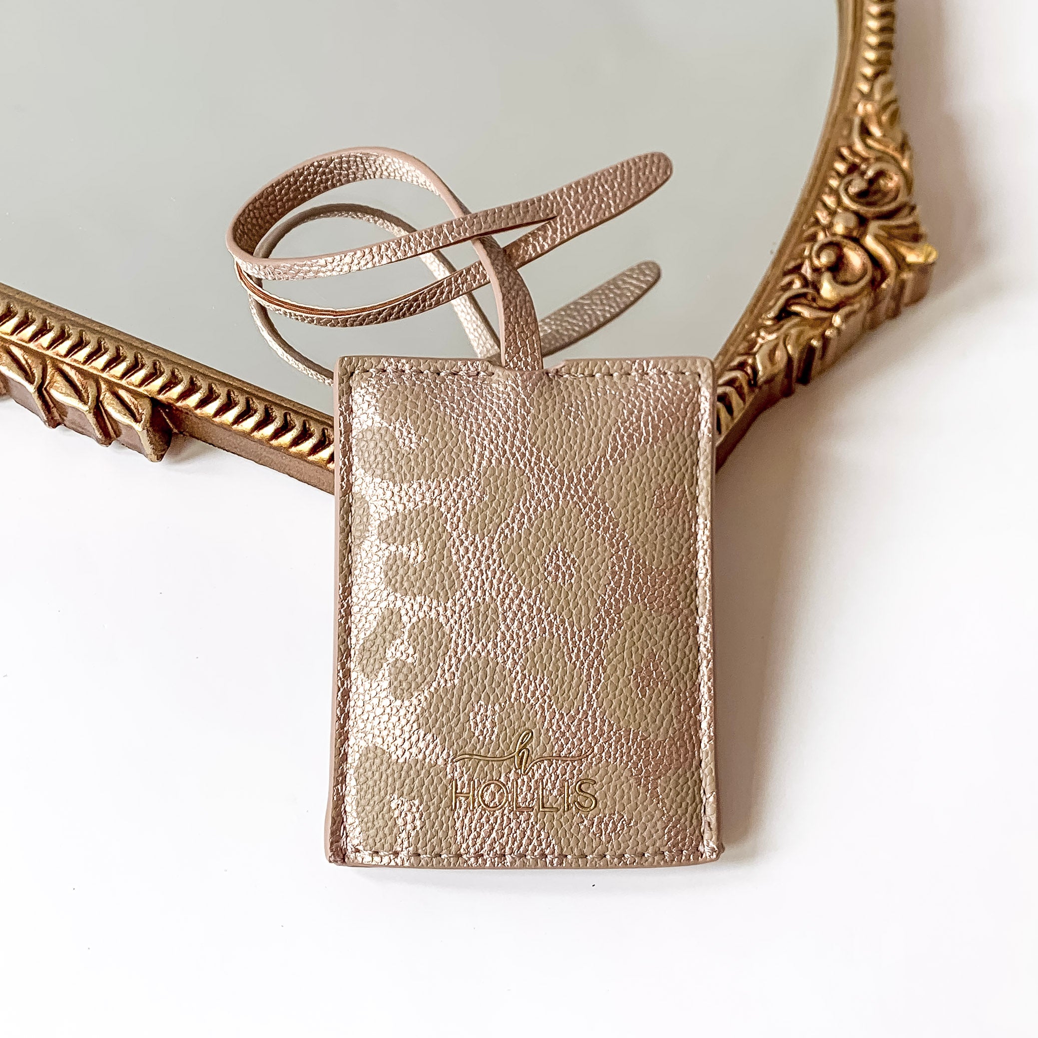 Leopard print luggage tag with a gold HOLLIS emblem at the bottom. This is pictured laying partially on a gold mirror on a white background.