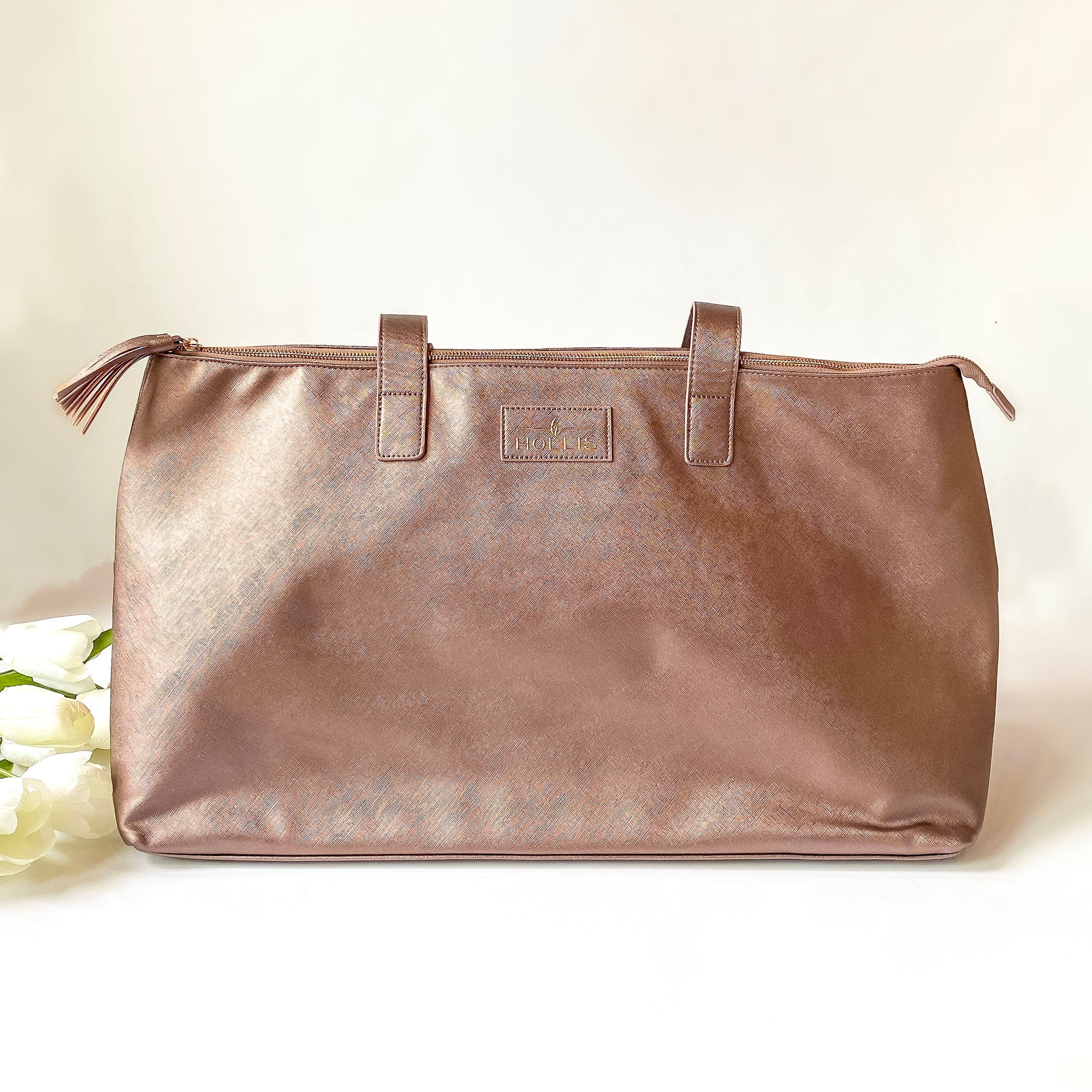 Metallic mocha duffle bag with a top zipper and metallic mocha handles. This bag is pictured on a white background with white flowers on the left side of the bag. 