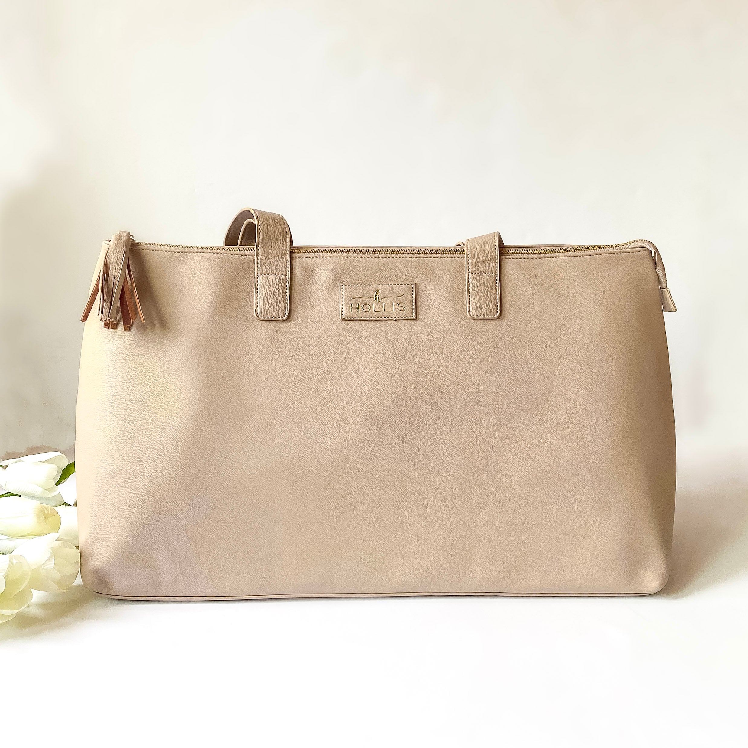 Nude duffle bag with a top zipper and nude handles. This bag is pictured on a white background with white flowers on the left side of the bag.