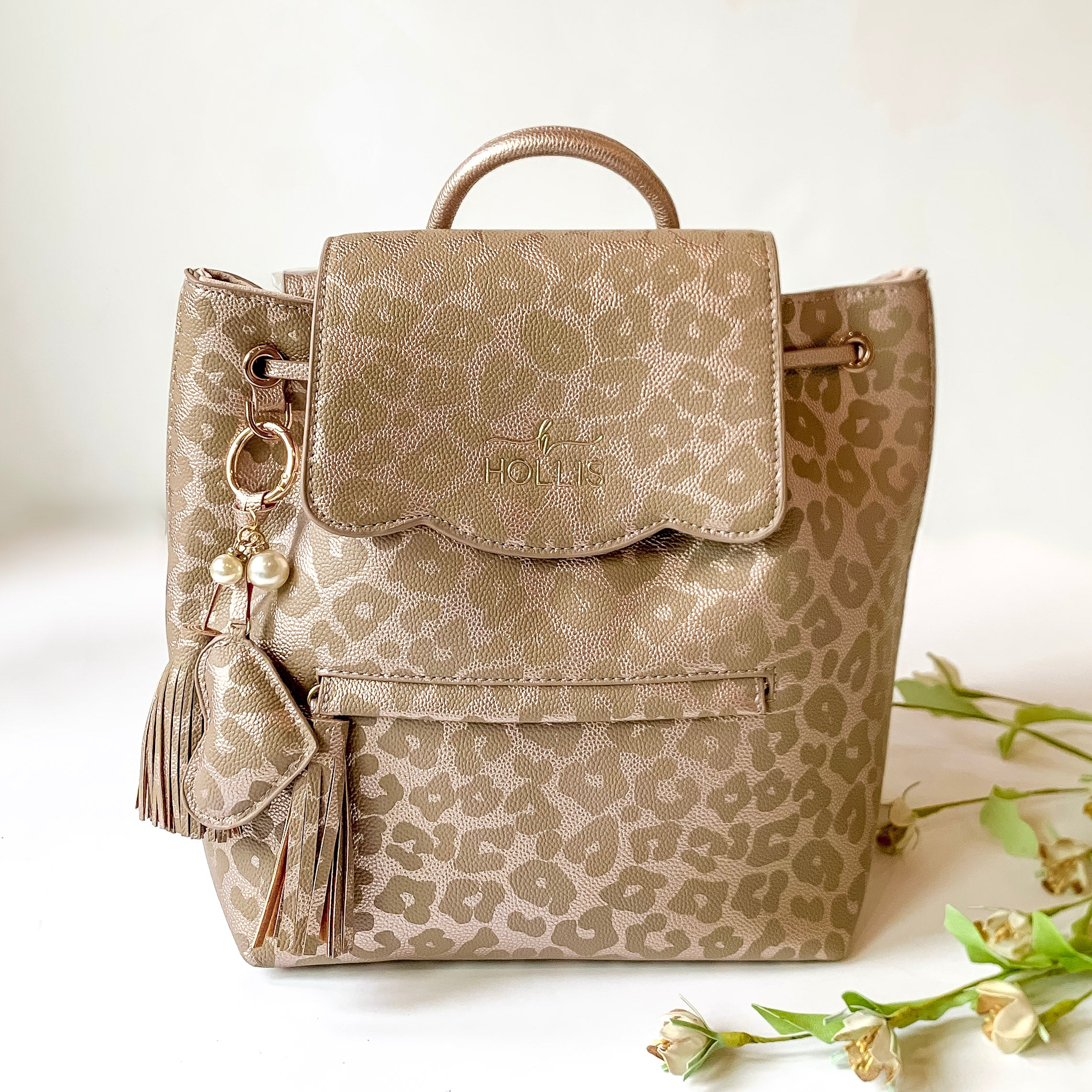 Leopard print backpack with a front zipper and front flap. This backpack also has a heart and tassel keychain. This backpack is pictured on a white background with flowers on the right side of the backpack. 