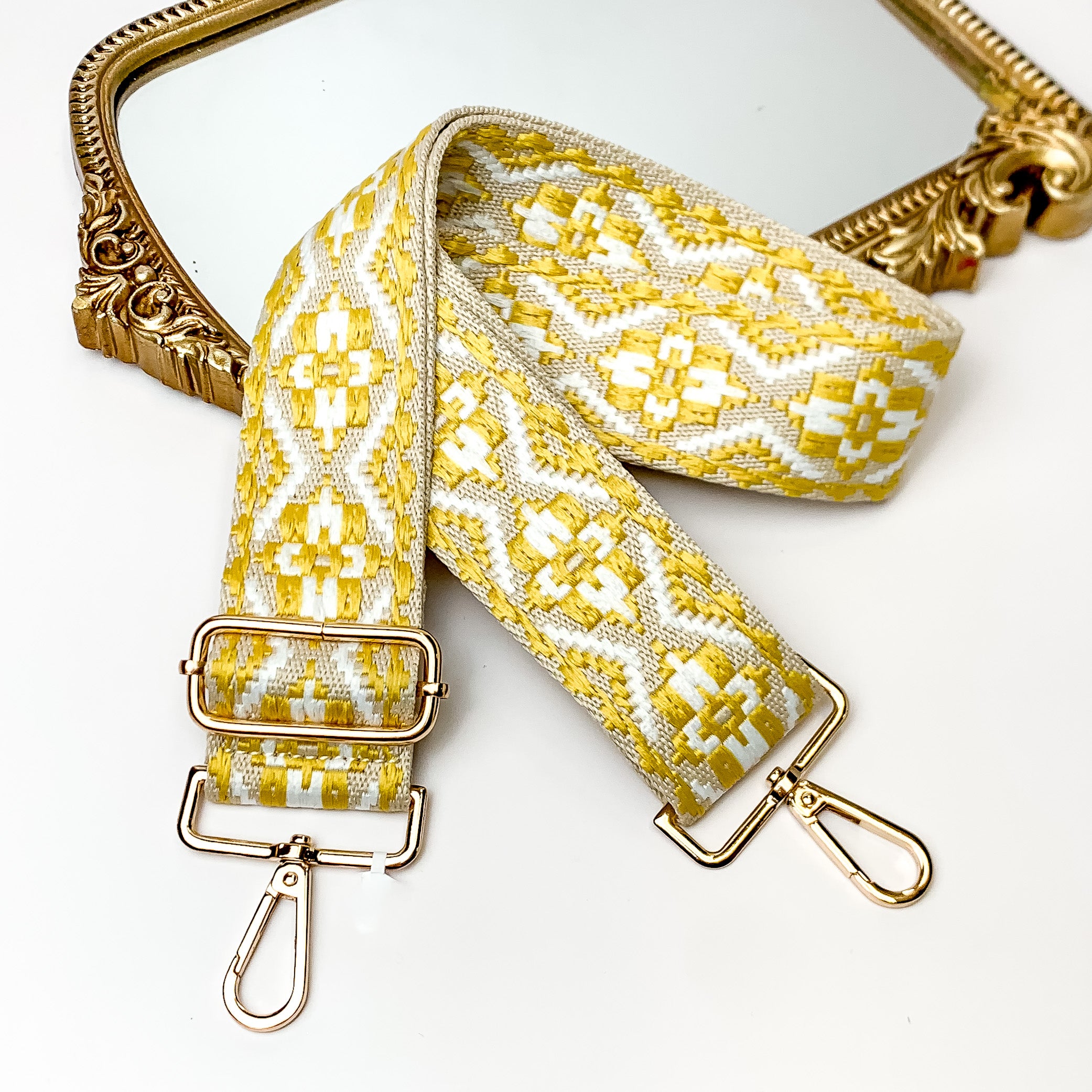 Beige canvas purse strap with white and yelow embroidered aztec design. This purse strap includes gold accents. This purse strap is pictured partially laying on a gold mirror on a white background.