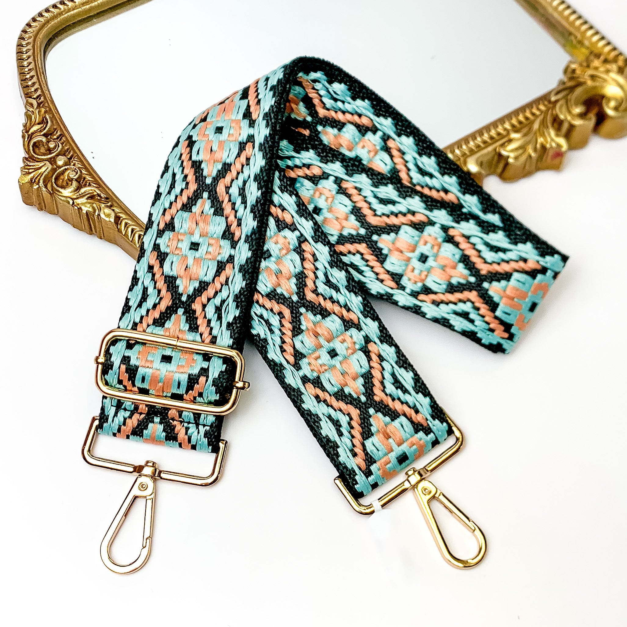 Black canvas purse strap with turquoise and peach embroidered aztec design. This purse strap includes gold accents. This purse strap is pictured partially laying on a gold mirror on a white background.