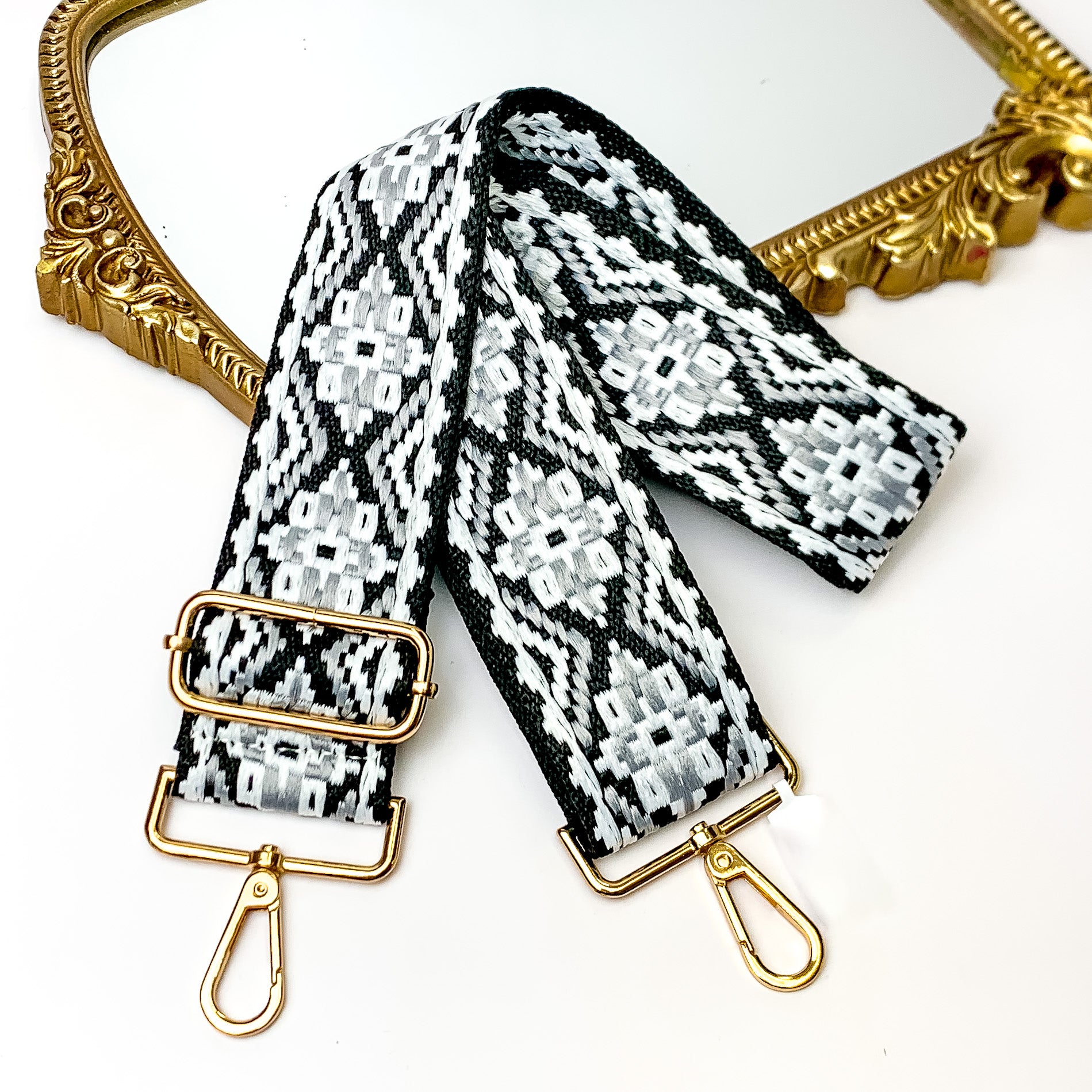 Black canvas purse strap with black and white embroidered aztec design. This purse strap includes gold accents. This purse strap is pictured partially laying on a gold mirror on a white background.