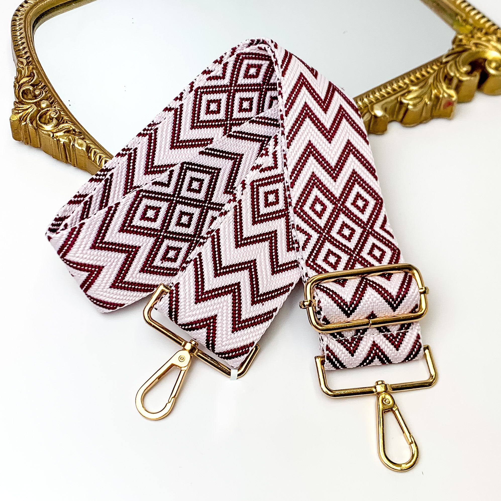Light pink canvas purse strap with maroon chevron and diamond design. This purse strap includes gold accents. This purse strap is pictured partially on a gold mirror on a white background.