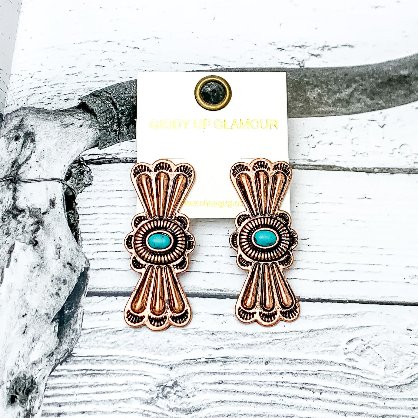 Lucky Break Hour-Glass Shaped Earrings in Copper Tone with Faux Turquoise Stone. Pictured on a wood like background with a longhorn on it.