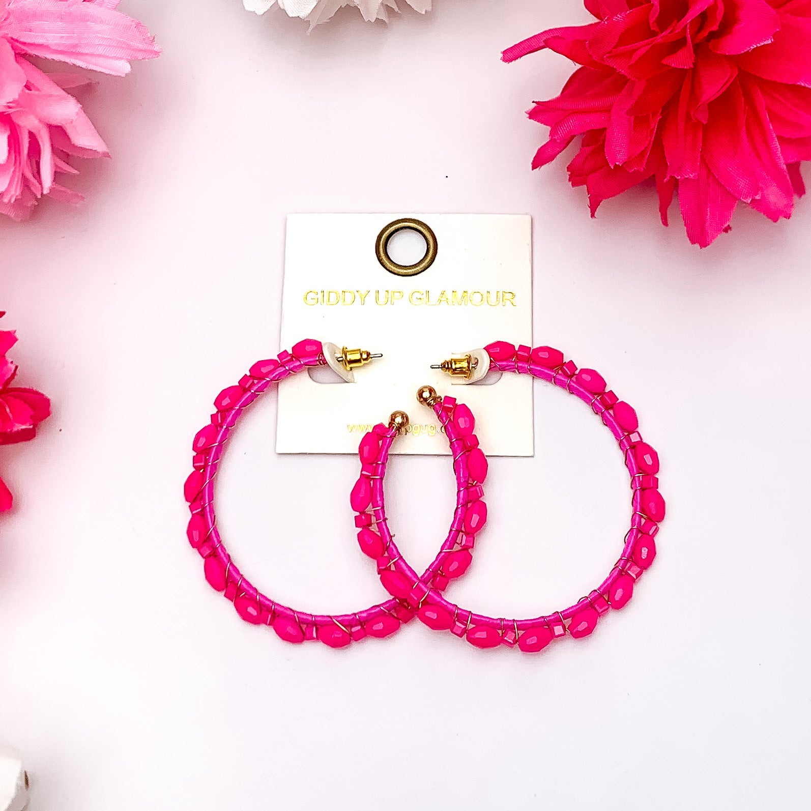Large Hoop Earrings Outlined with Crystals in Hot Pink. Pictured on a white background surrounded by pink flowers.
