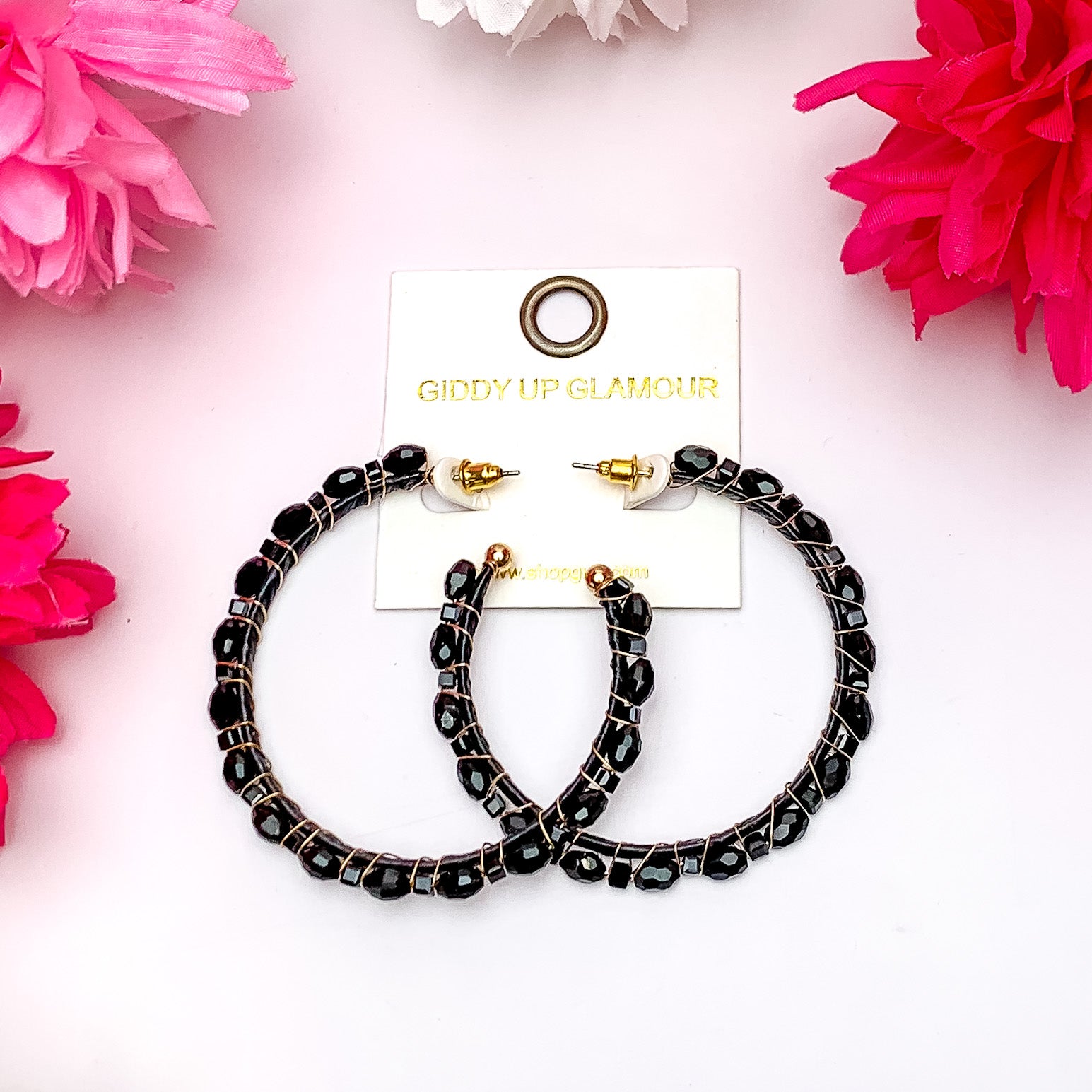 Large Hoop Earrings Outlined with Crystals in Black. Pictured on a white background surrounded by pink flowers.