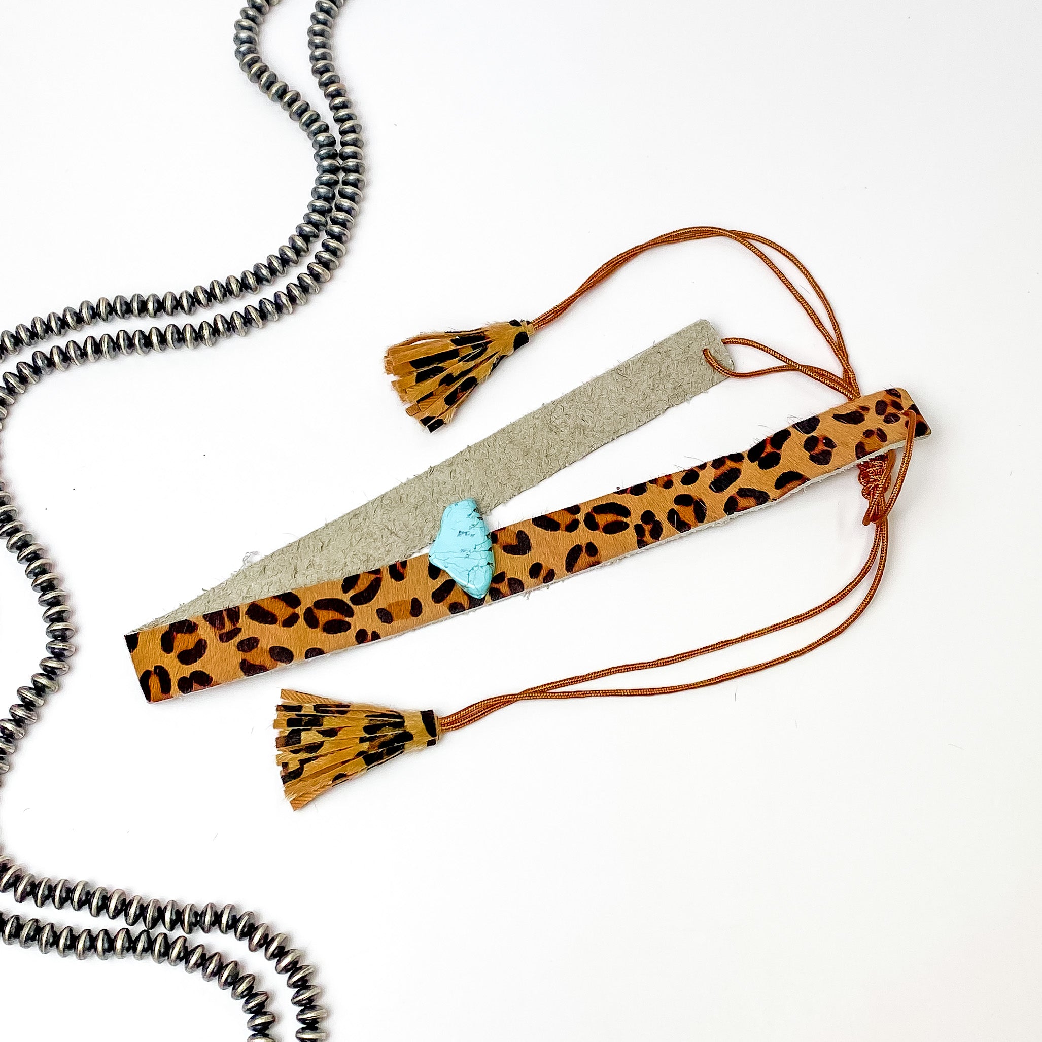 Pictured is a leopard print, adjustable hat band with a turquoise stone. This hat band is pictured on a white background with silver, saucer beads on the left side of the hat band. 