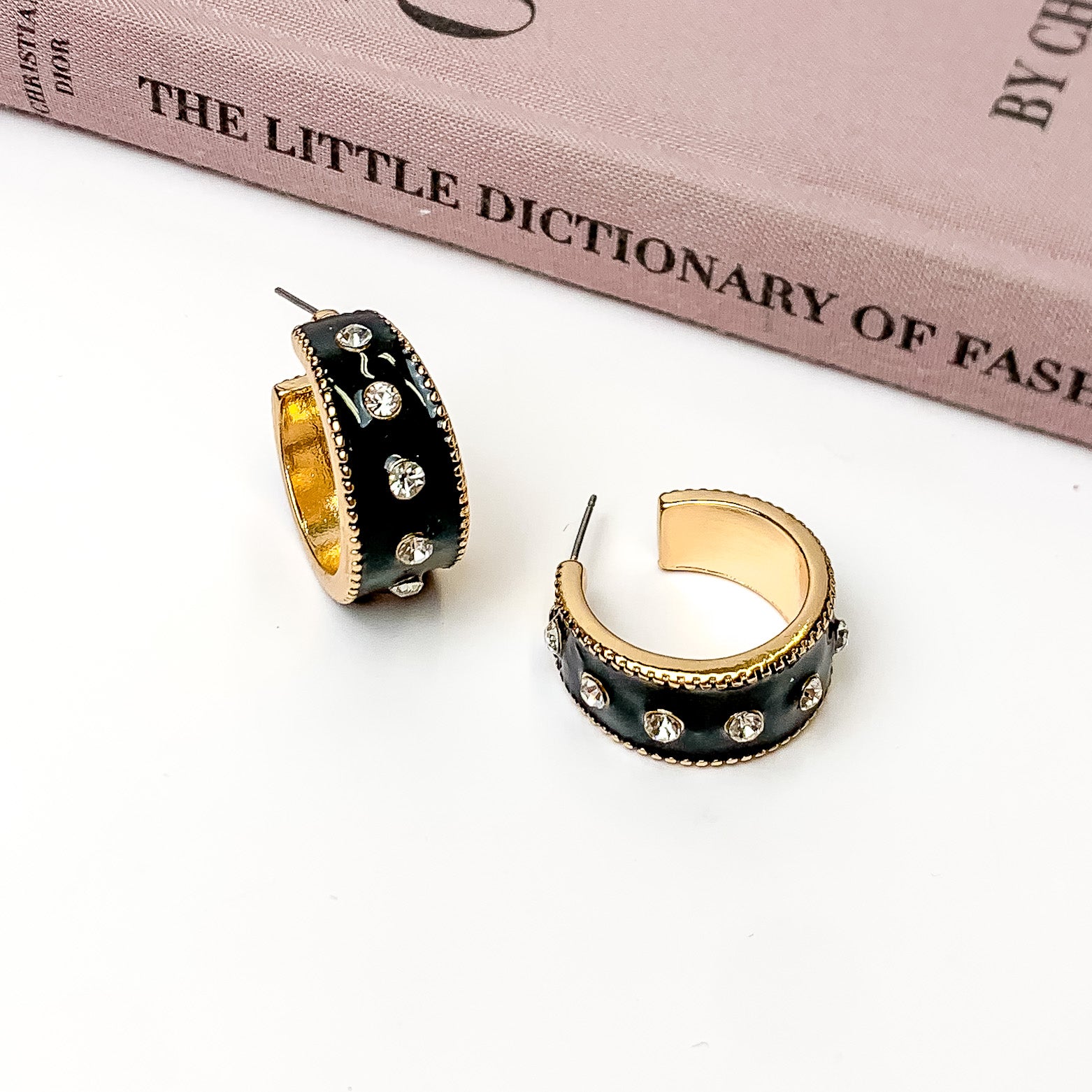 Thick, gold hoop earrings with a black inlay on the outside. These hoops also include a single line of clear crystals through the middle. One hoop is pictured stadning up right and the other is pictured laying on its side. These hoops are pictured on a white background with a mauve colored book above the earrings. 
