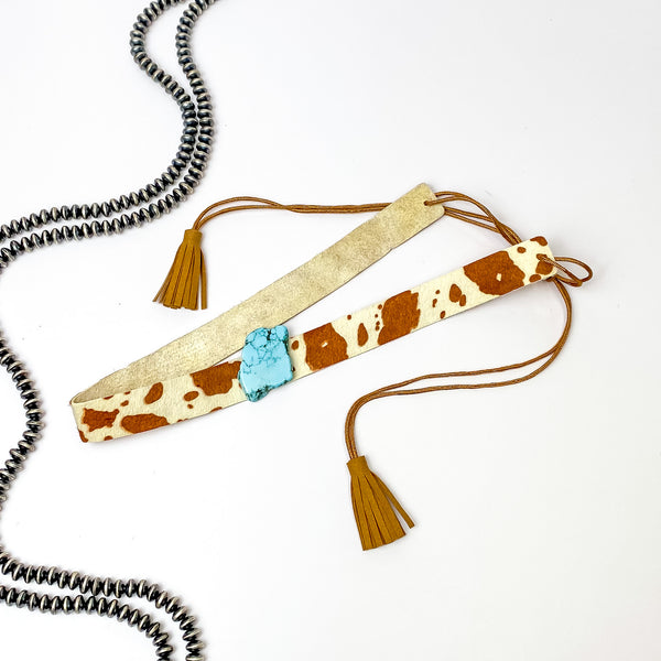Pictured is a cow print, adjustable hat band with a turquoise stone charm. This hat band is pictured on a white background with silver, saucer beads on the left side of the hat band. 