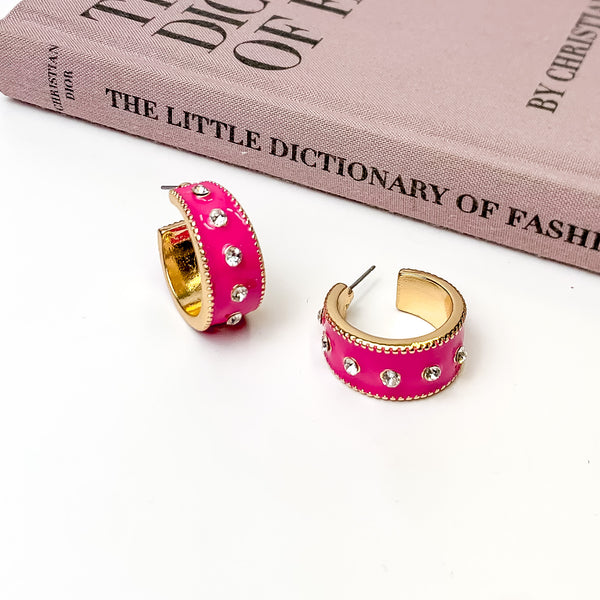 Thick, gold hoop earrings with a fuchsia pink inlay on the outside. These hoops also include a single line of clear crystals through the middle. One hoop is pictured stadning up right and the other is pictured laying on its side. These hoops are pictured on a white background with a mauve colored book above the earrings. 