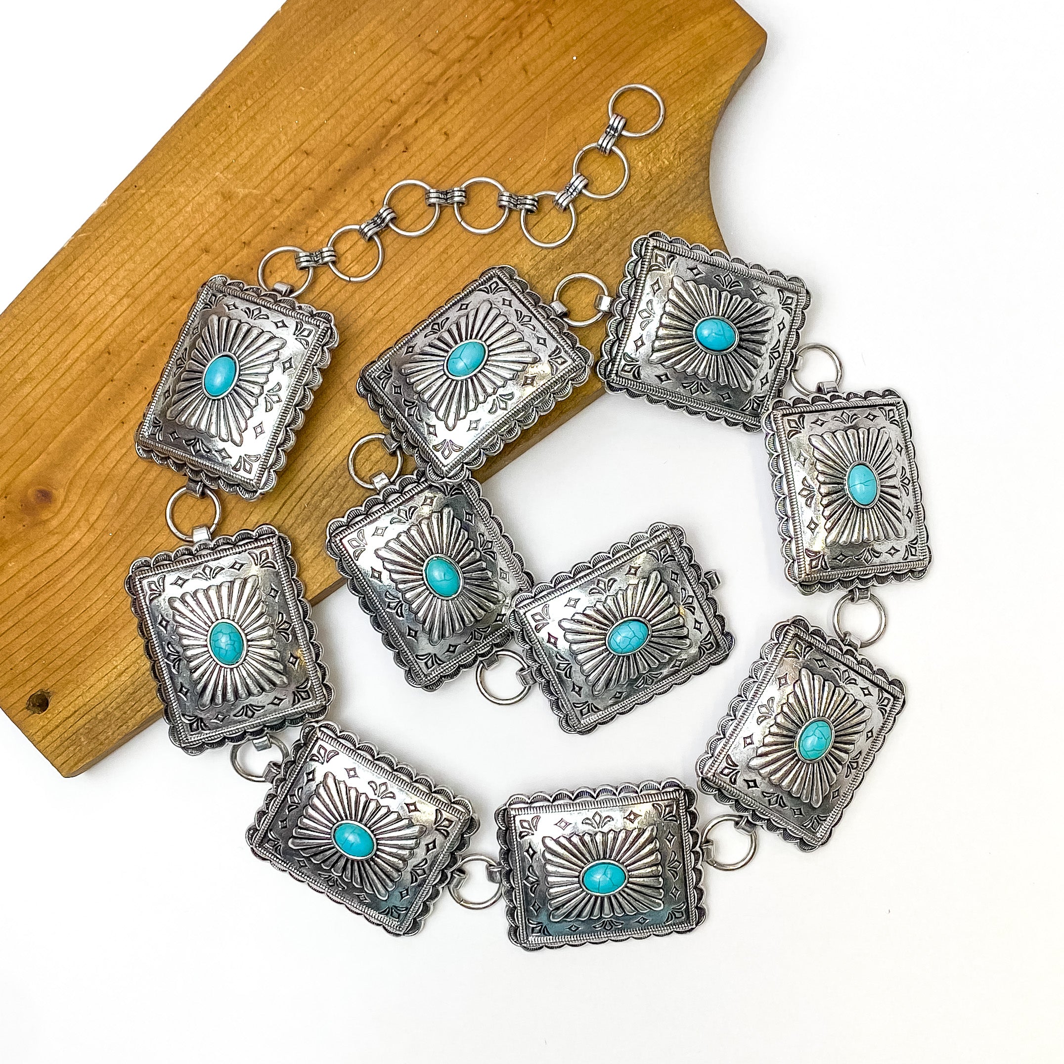 Pictured is a silver, square concho belt with detailing and turquoise center stones. This belt is pictured in a spiral on a white and brown background. 