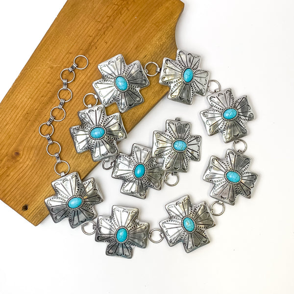 Pictured is a silver, cross concho belt with detailing and turquoise center stones. This belt is pictured in a spiral on a white and brown background. 