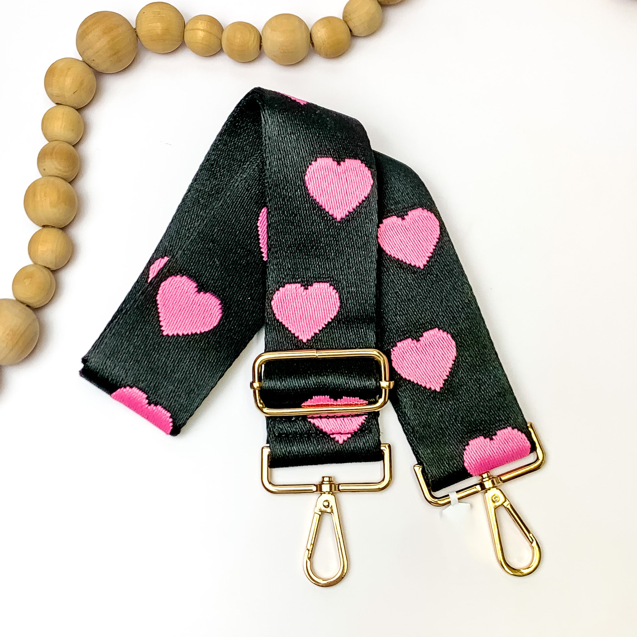 Black purse strap with a pink heart design. This purse strap includes gold hardware. This purse strap os pictured on a white background with tan beads in the top left corner. 