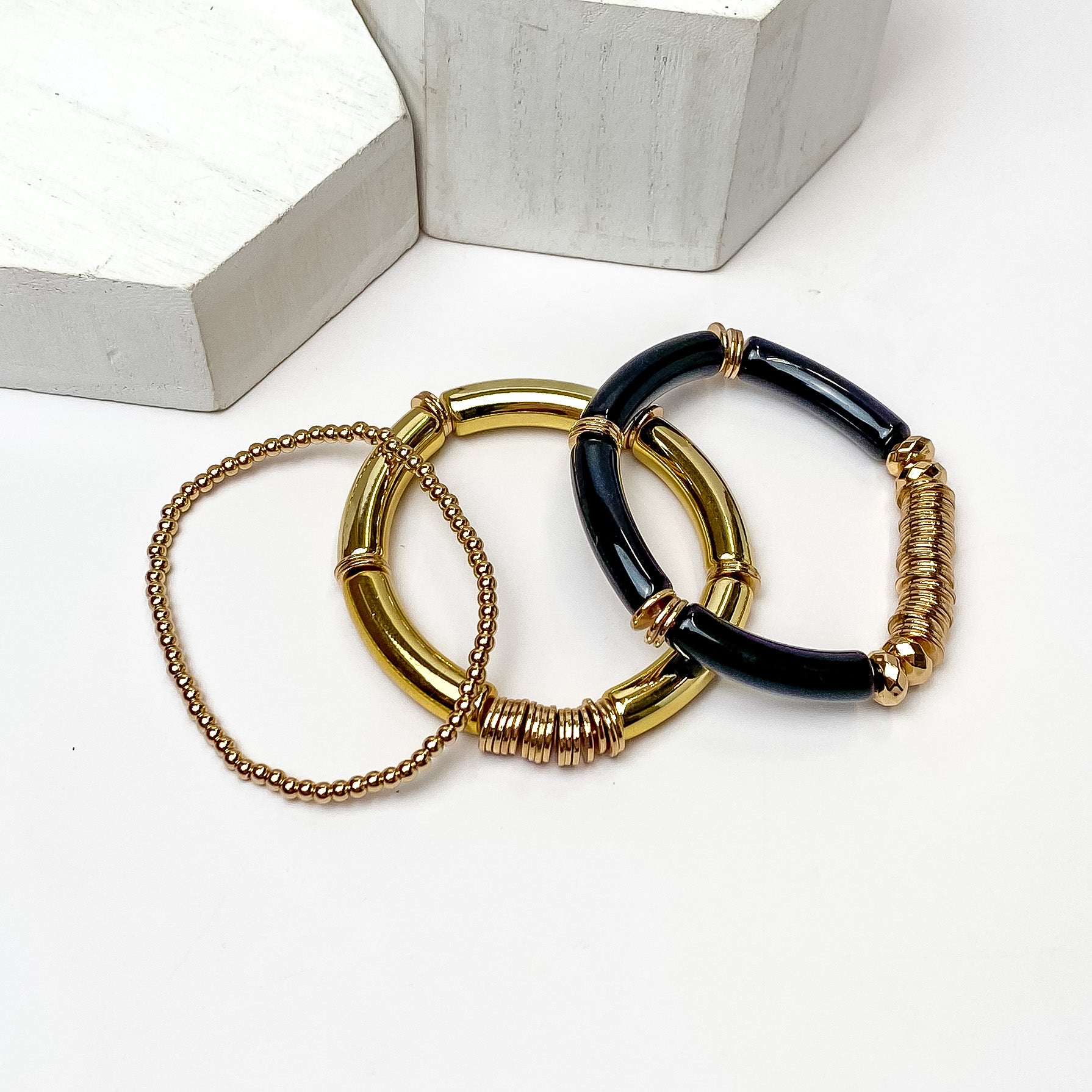 Set of Three | Bahama Nights Gold Tone Stretchy Bracelets With Accents of Black. Pictured on a white background with white objects above the bracelets. 