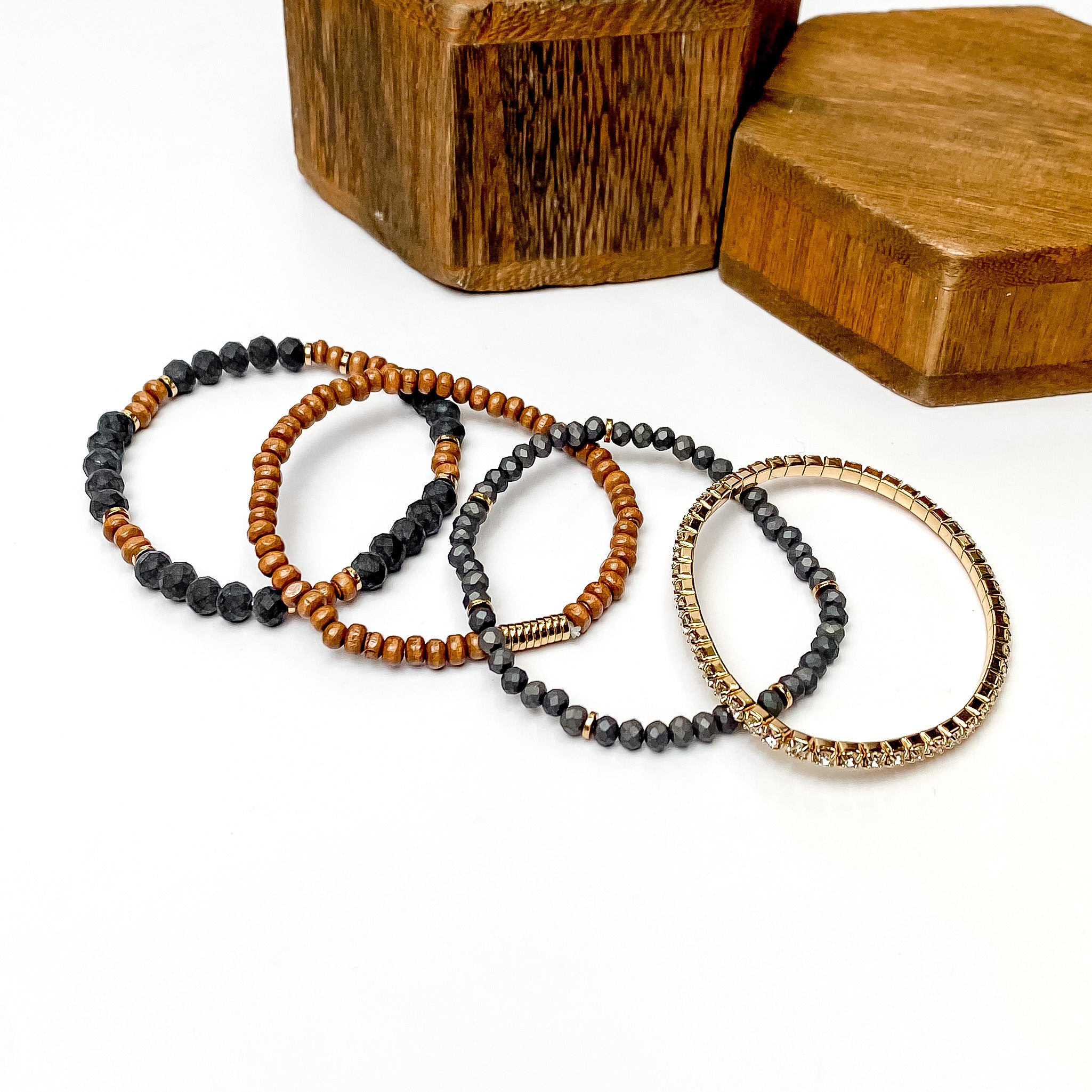 Set of four bracelets pictured laying one on top of the other. The bracelet at the top includes black and brown beads, the second has brown beads and gold disc beads, the third includes black beads with gold disk spacers, and the last bracelet is a full crystal band with a gold setting. These bracelets are pictured on a white background with brown blocks in the top right corner. 