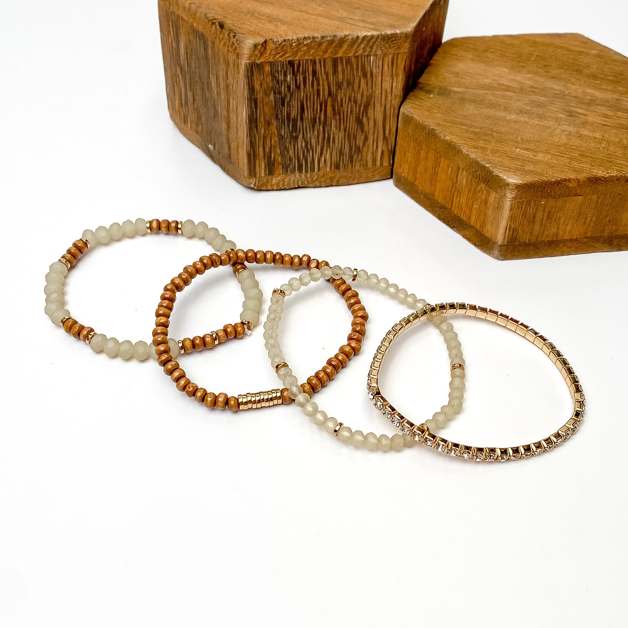 Set of four bracelets pictured laying one on top of the other. The bracelet at the top includes white and brown beads, the second has brown beads and gold disc beads, the third includes ivory beads with gold disk spacers, and the last bracelet is a full crystal band with a gold setting. These bracelets are pictured on a white background with brown blocks in the top right corner. 