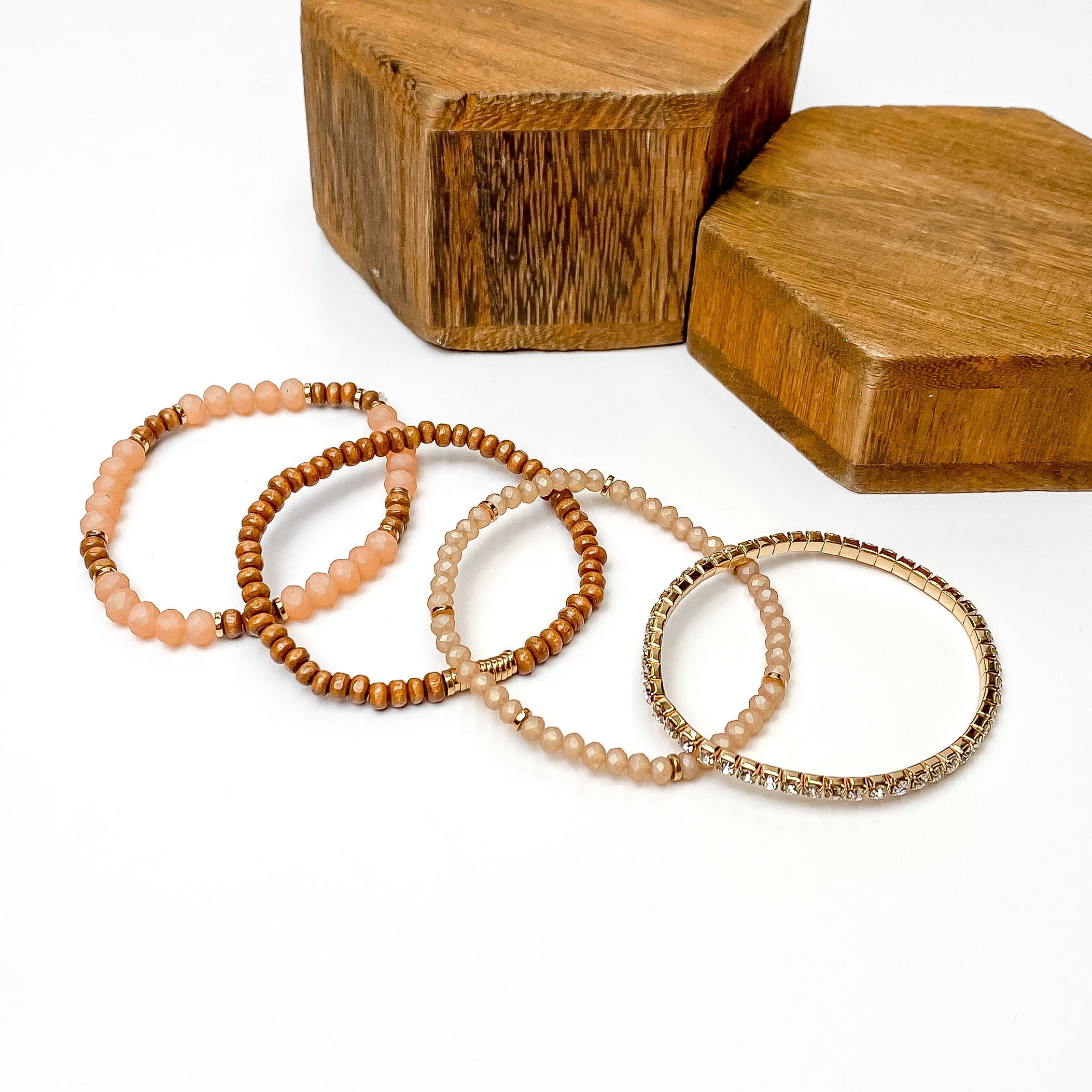 Set of four bracelets pictured laying one on top of the other. The bracelet at the top includes dusty pink and brown beads, the second has brown beads and gold disc beads, the third includes dusty pink beads with gold disk spacers, and the last bracelet is a full crystal band with a gold setting. These bracelets are pictured on a white background with brown blocks in the top right corner. 
