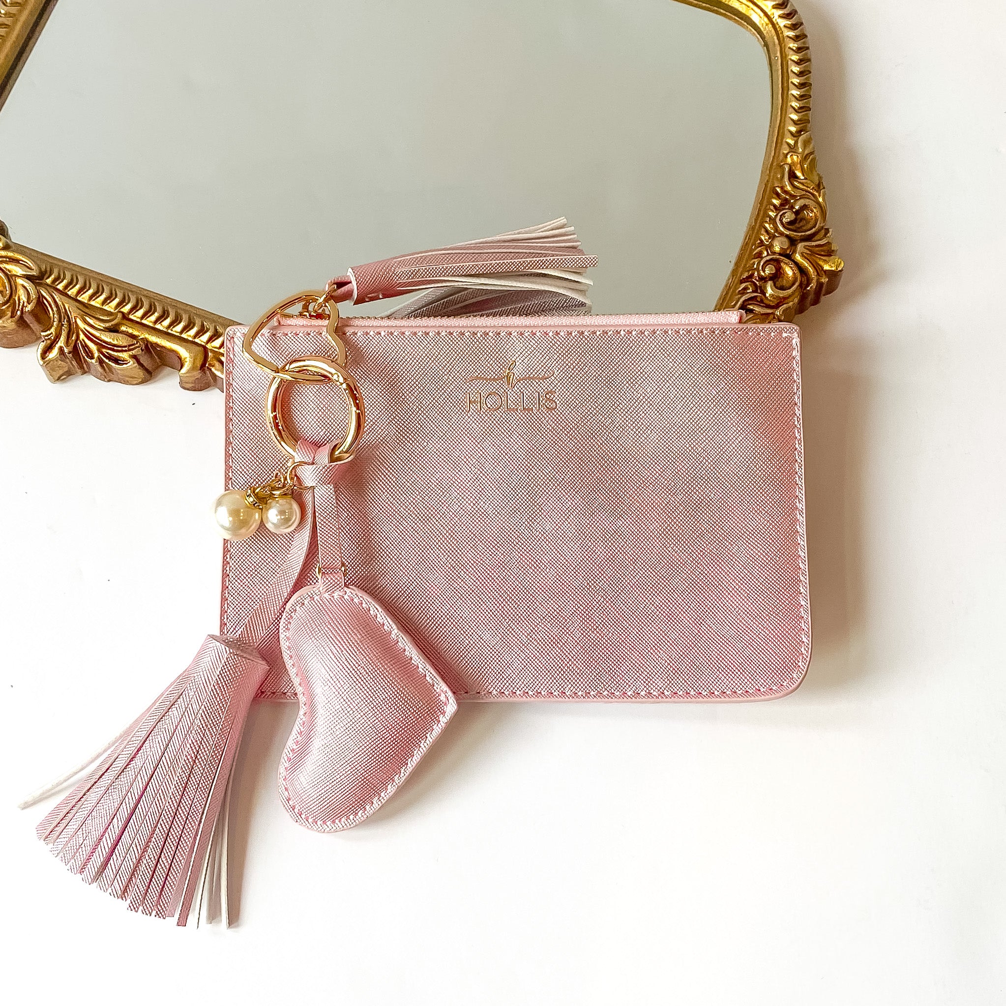 Metallic blush pink coin pouch with a gold HOLLIS emblem at the top and a top zipper. This coin pouch has a gold key ring, gold heart key ring, a blush tassel charm, a blush heart charm and white pearl charms. This luggage tag is pictured partially laying on a gold mirror on a white background. 