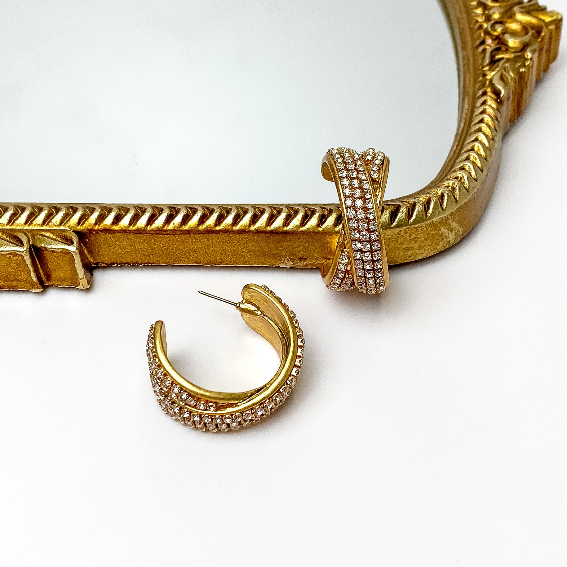 Two gold bands that are covered in clear crystals are crossed in the center and formed to make hoop earrings. These earrings are pictured on a white background with a gold mirror at the top of the picture. 